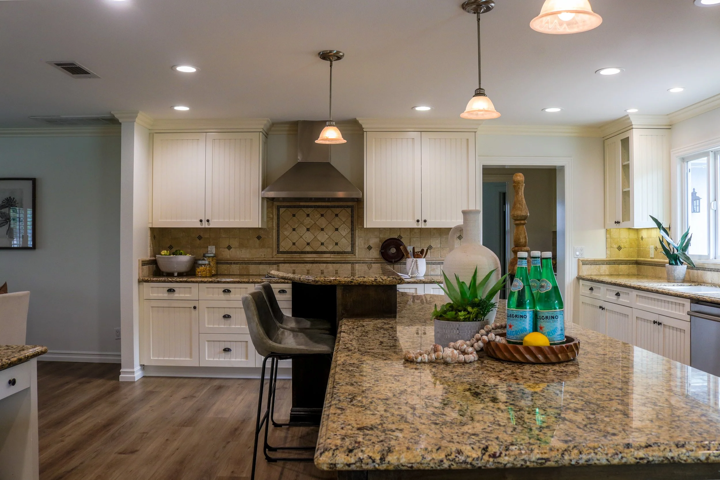 A spacious kitchen with white cabinetry, a granite island, and pendant lights hanging from the ceiling. There are bottles of sparkling water and decorative plants on the counter, with a window letting in natural light.