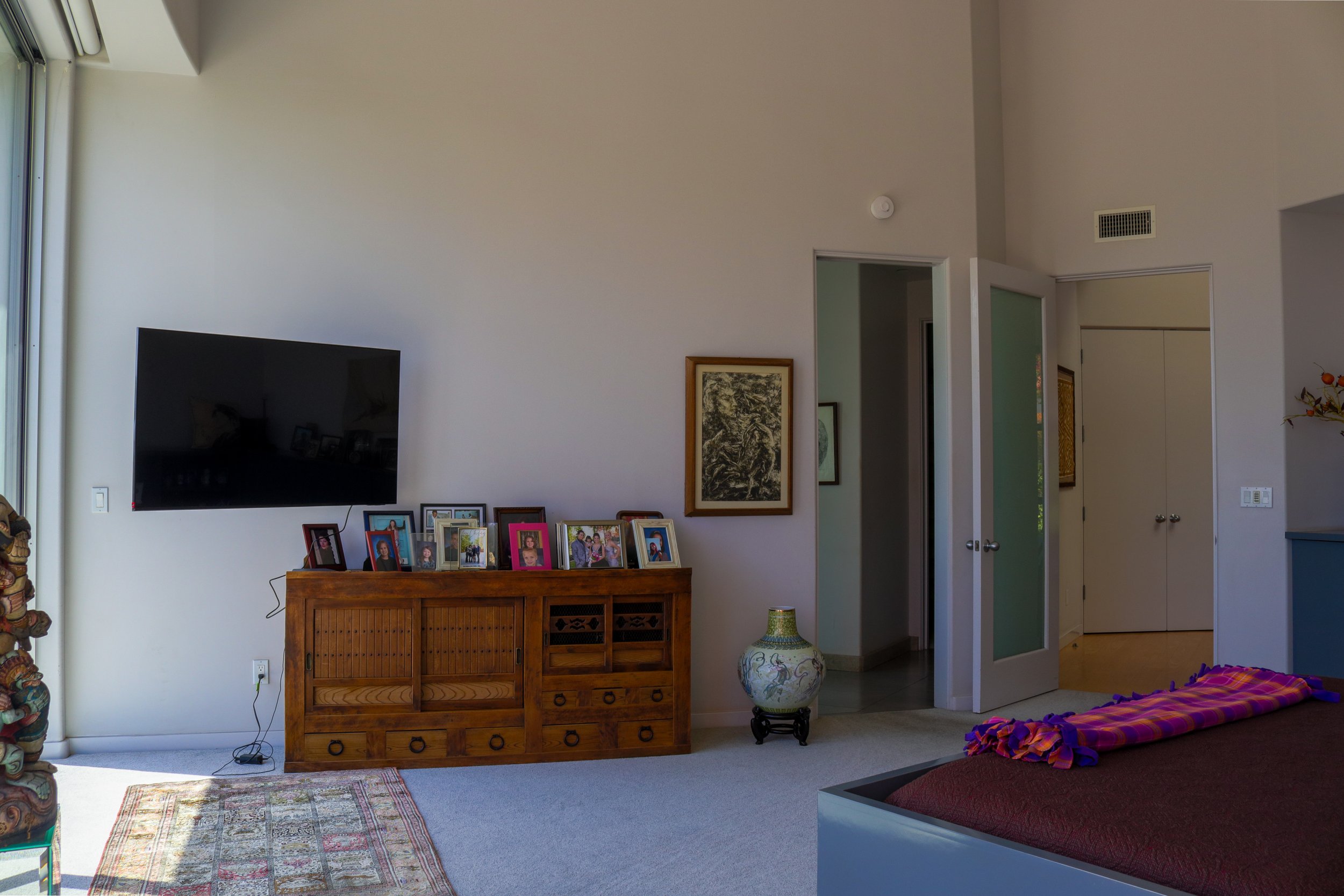 A living room with a mounted flat-screen TV on a white wall. Below it, a wooden sideboard with framed photos. There is a patterned rug on the beige carpet floor. A large decorative vase is on a small black stand between an open doorway and a closed c