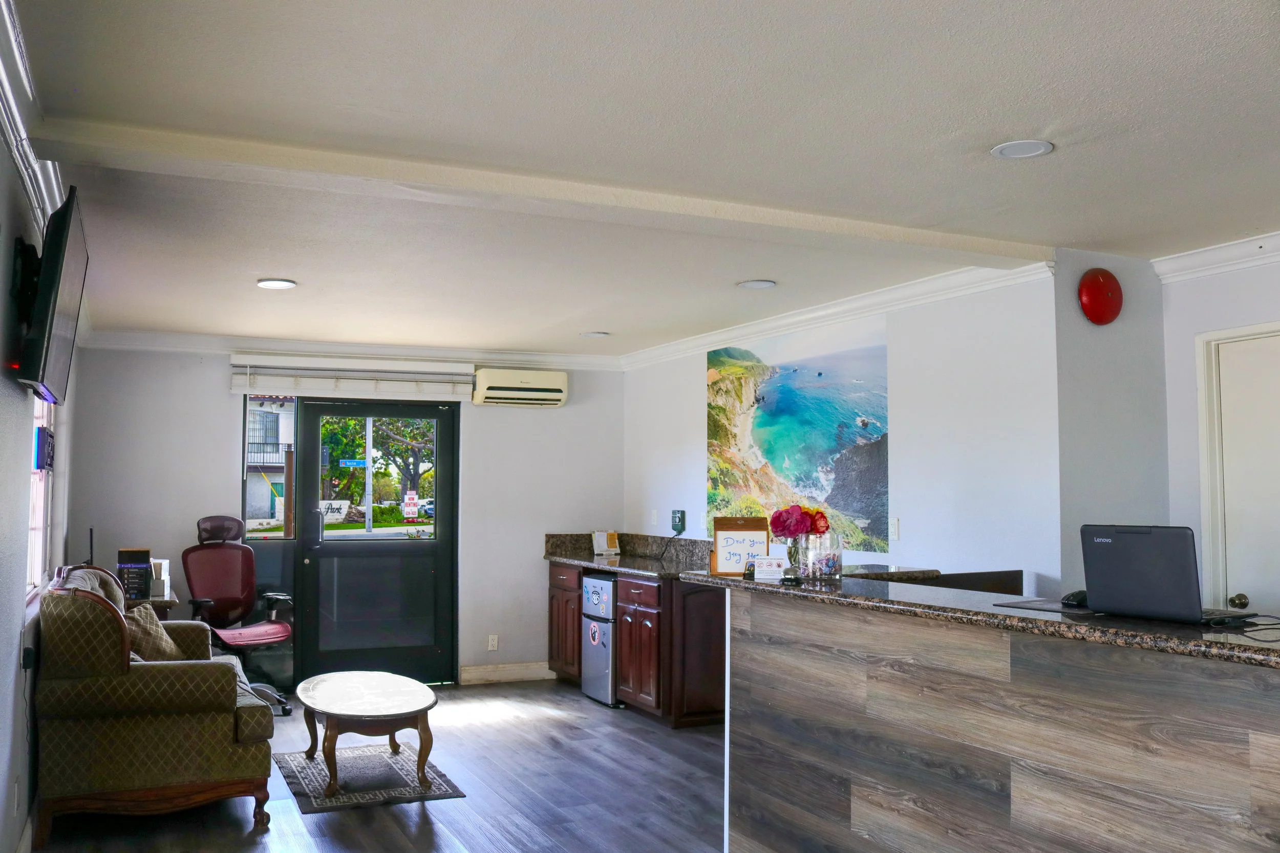 Interior of a reception area with a seating area, a kitchen counter, and a door leading outdoors, decorated with a large scenic beach photo on the wall.