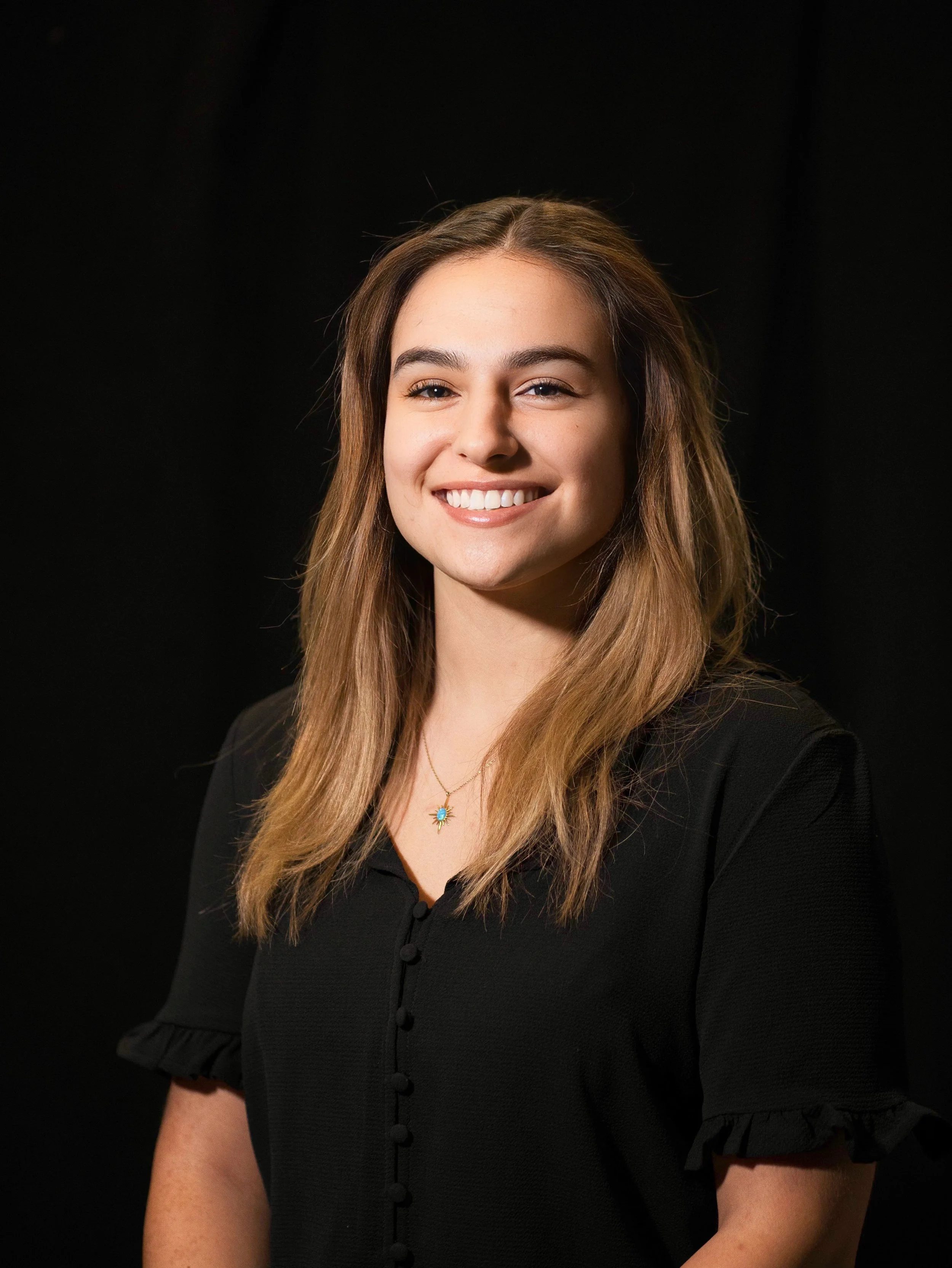 Portrait of a young woman with light brown hair, smiling, wearing a black top and a necklace with a blue pendant, against a black background.