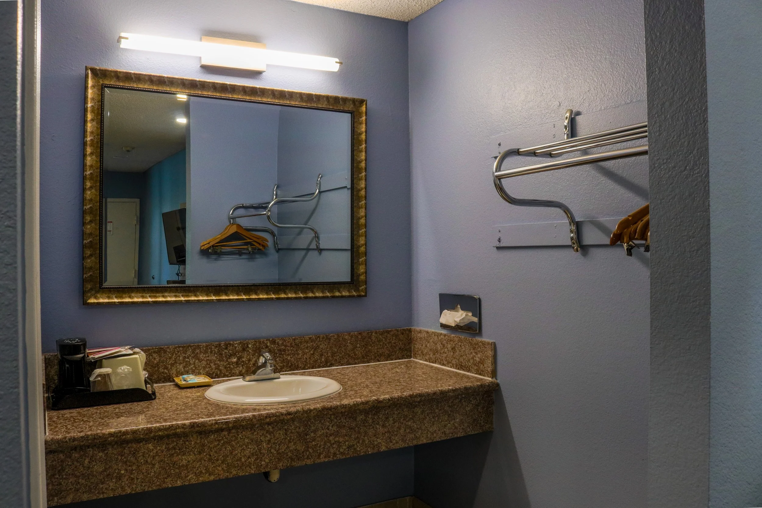 A bathroom sink counter with a mirror above, washing supplies, a tissue box, and a wall-mounted towel rack with hangers.