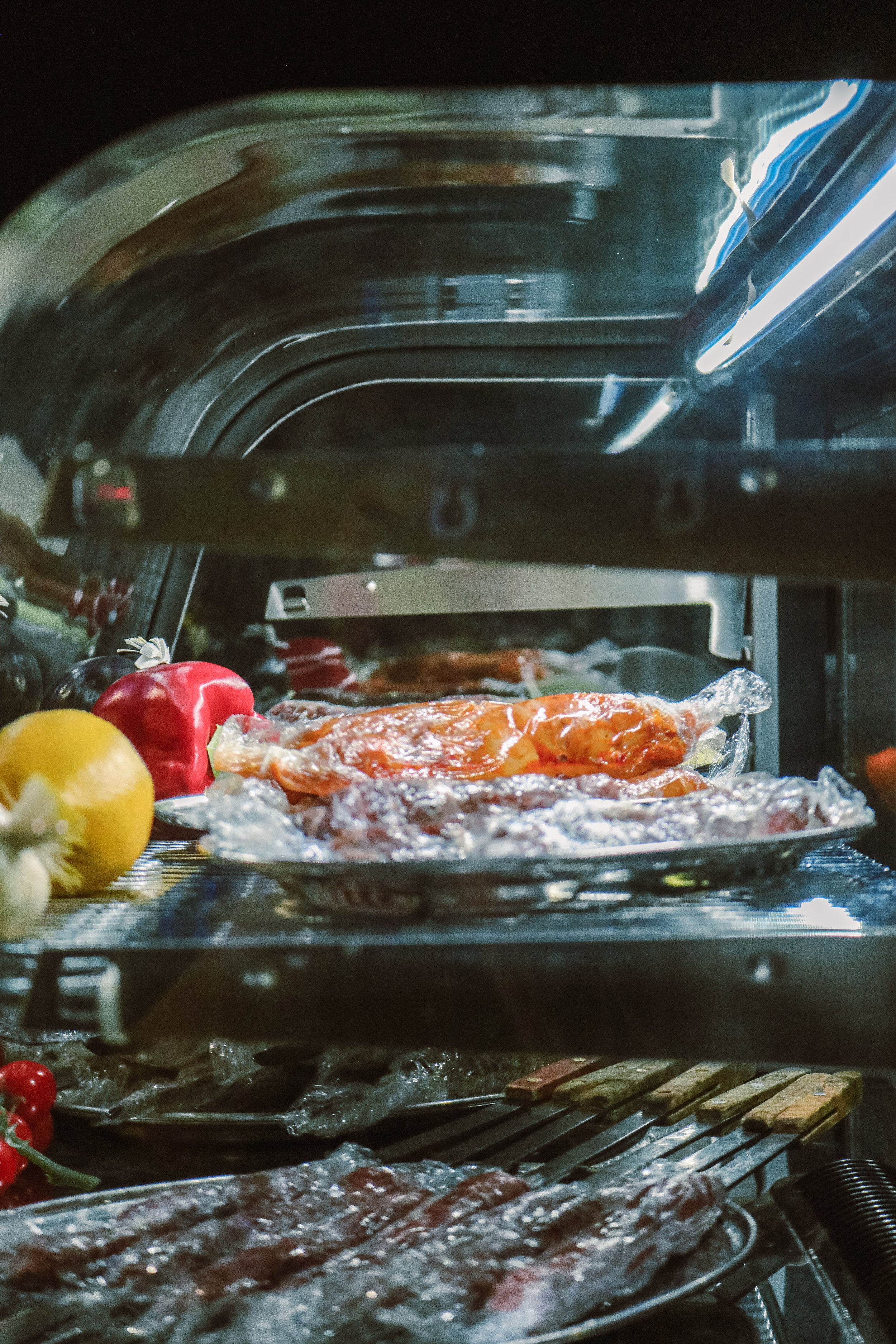 Inside a refrigerator with food wrapped in aluminum foil, including strips of meat, and fresh strawberries and lemons in the foreground.