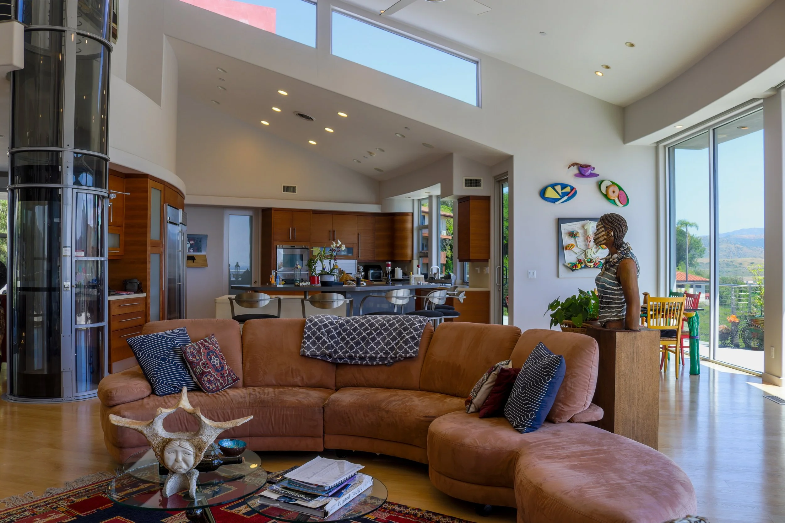 Open living room with a brown sectional sofa, a coffee table with magazines and decorative items, a kitchen with wooden cabinets and a bar area in the background, large windows letting in natural light, colorful wall art, and a woman standing near th