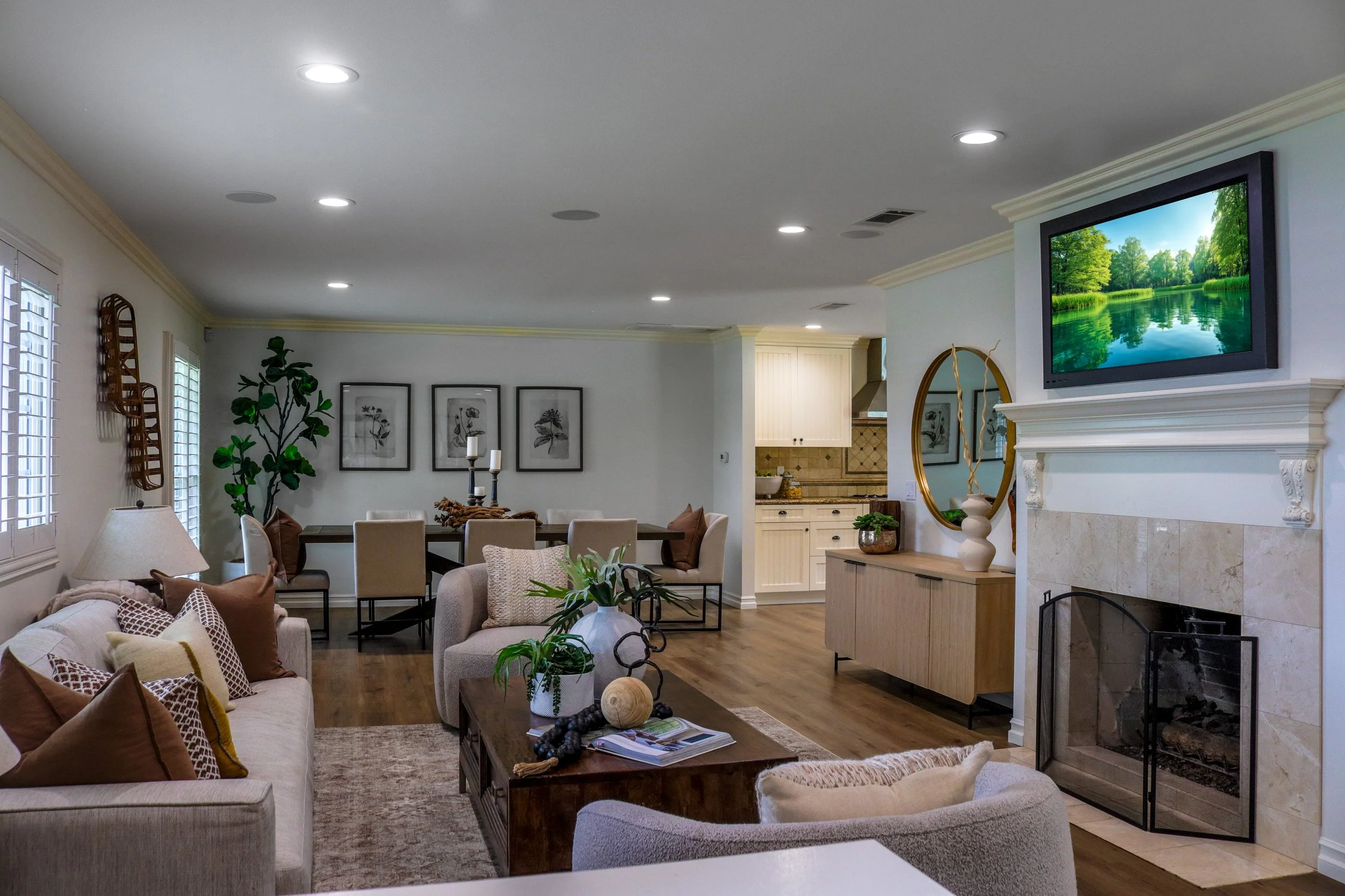 Living room with a fireplace, flat-screen TV, dining table, and various decorative items, with windows and artwork on the walls.