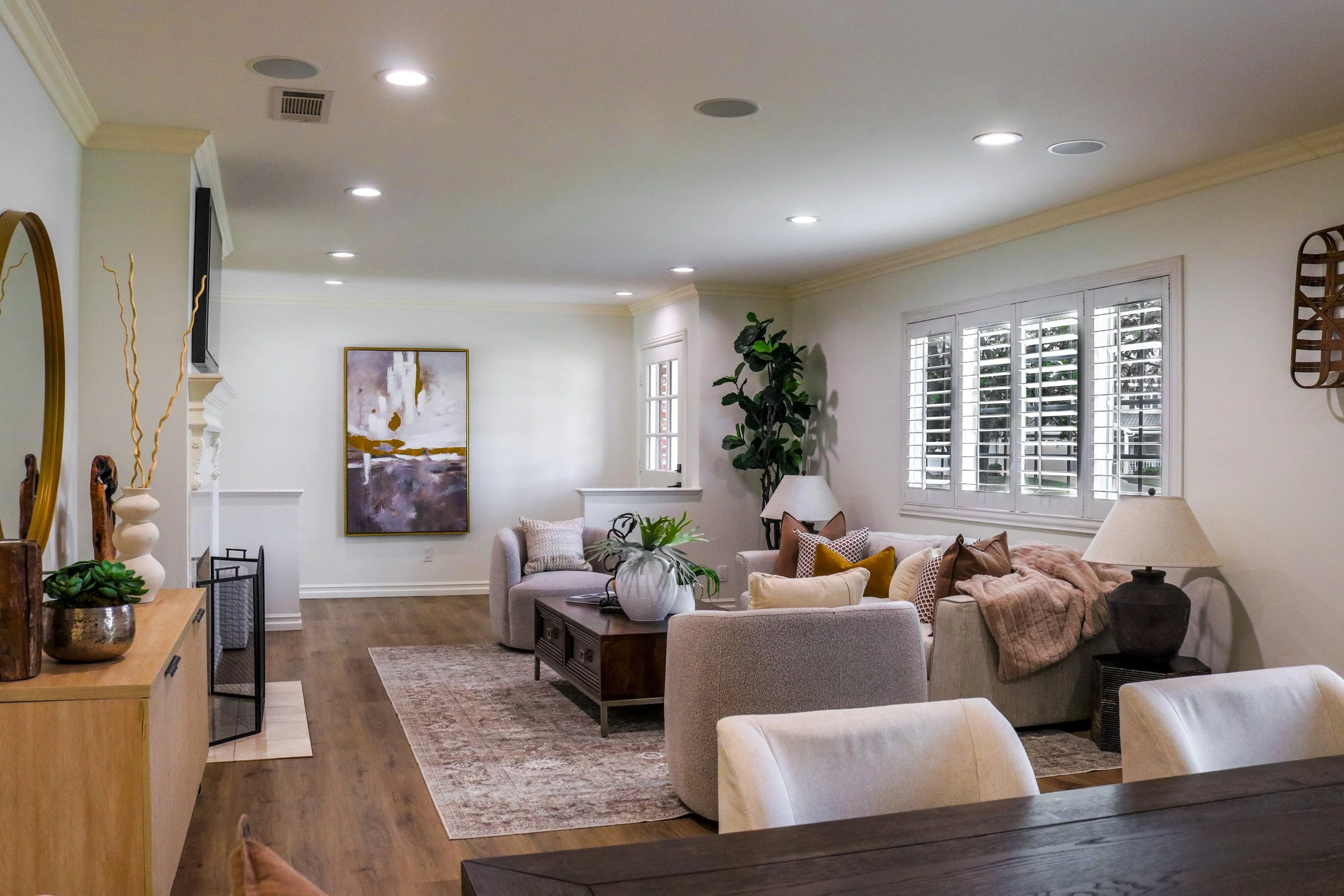 Living room with white walls, large windows with shutters, ceiling lights, beige and white furniture, artwork on the wall, and various decorative plants and items.