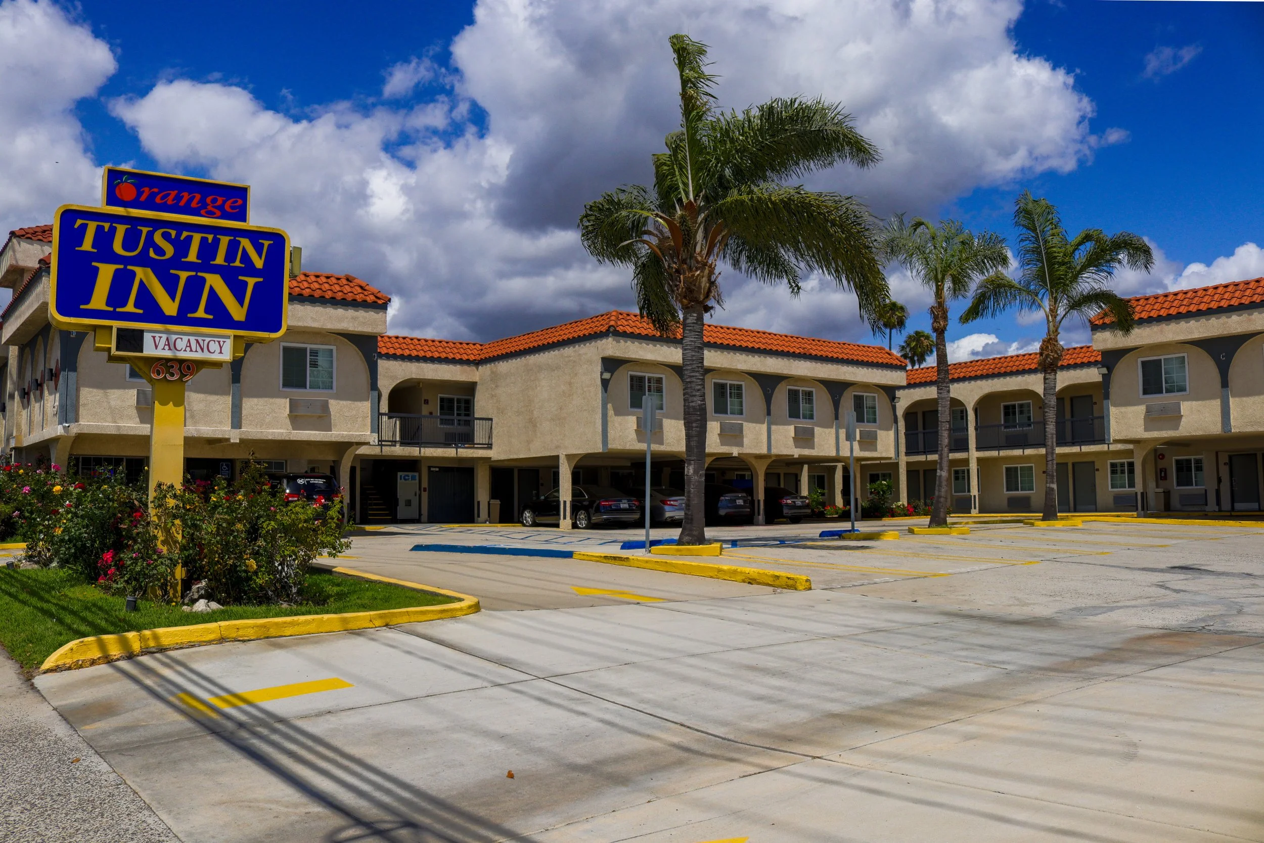 The image shows a hotel parking lot with a sign for the Tustin Inn, part of the Orange hotel chain, with a vacancy sign. The building has a stucco exterior with red tile roofing, and several palm trees are in front of it. The sky is partly cloudy wit