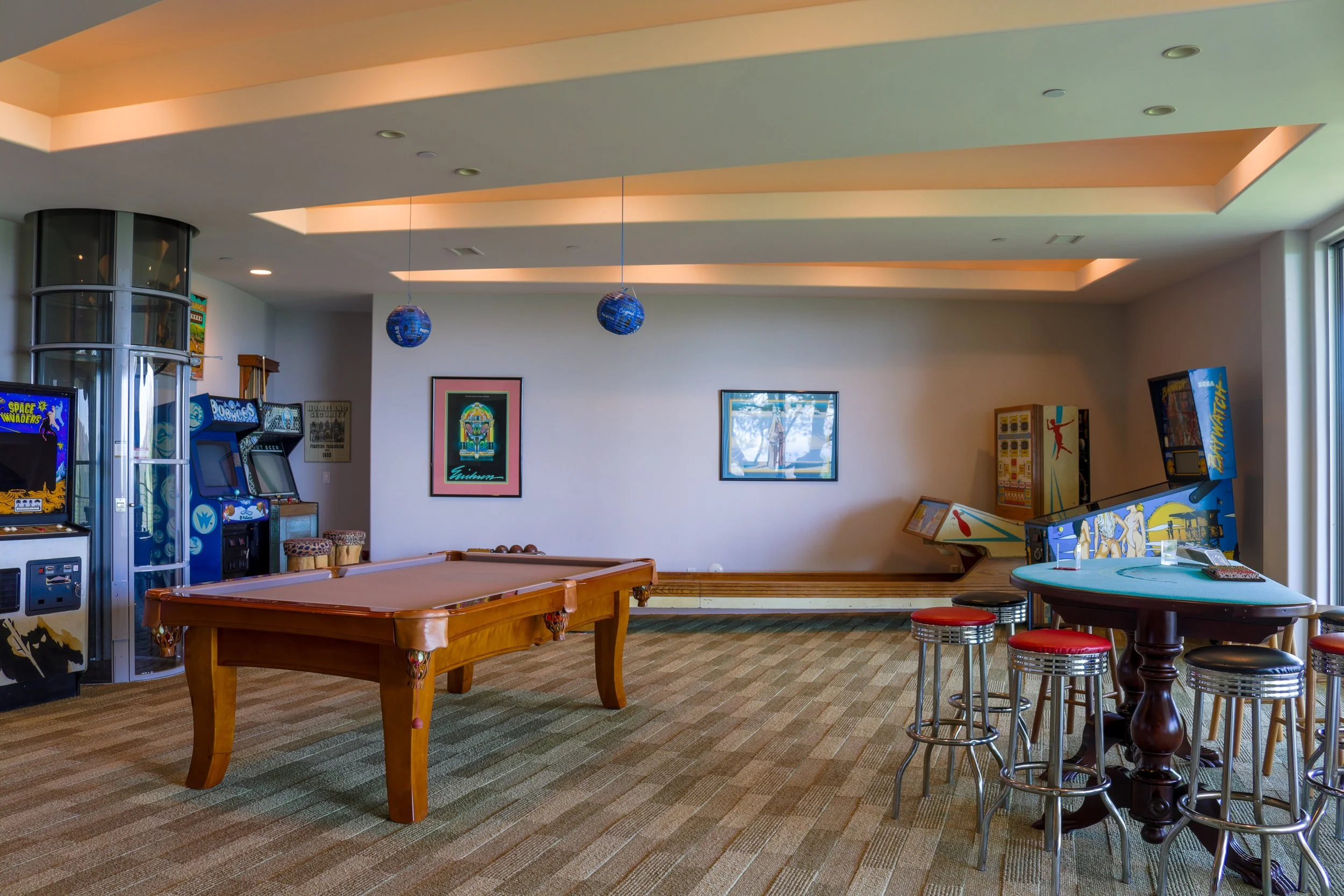 Indoor arcade room with vintage pinball machines, a pool table, and a round table with stools. Decor includes framed posters and blue globe hanging lights, with carpeted flooring and lounge chairs.