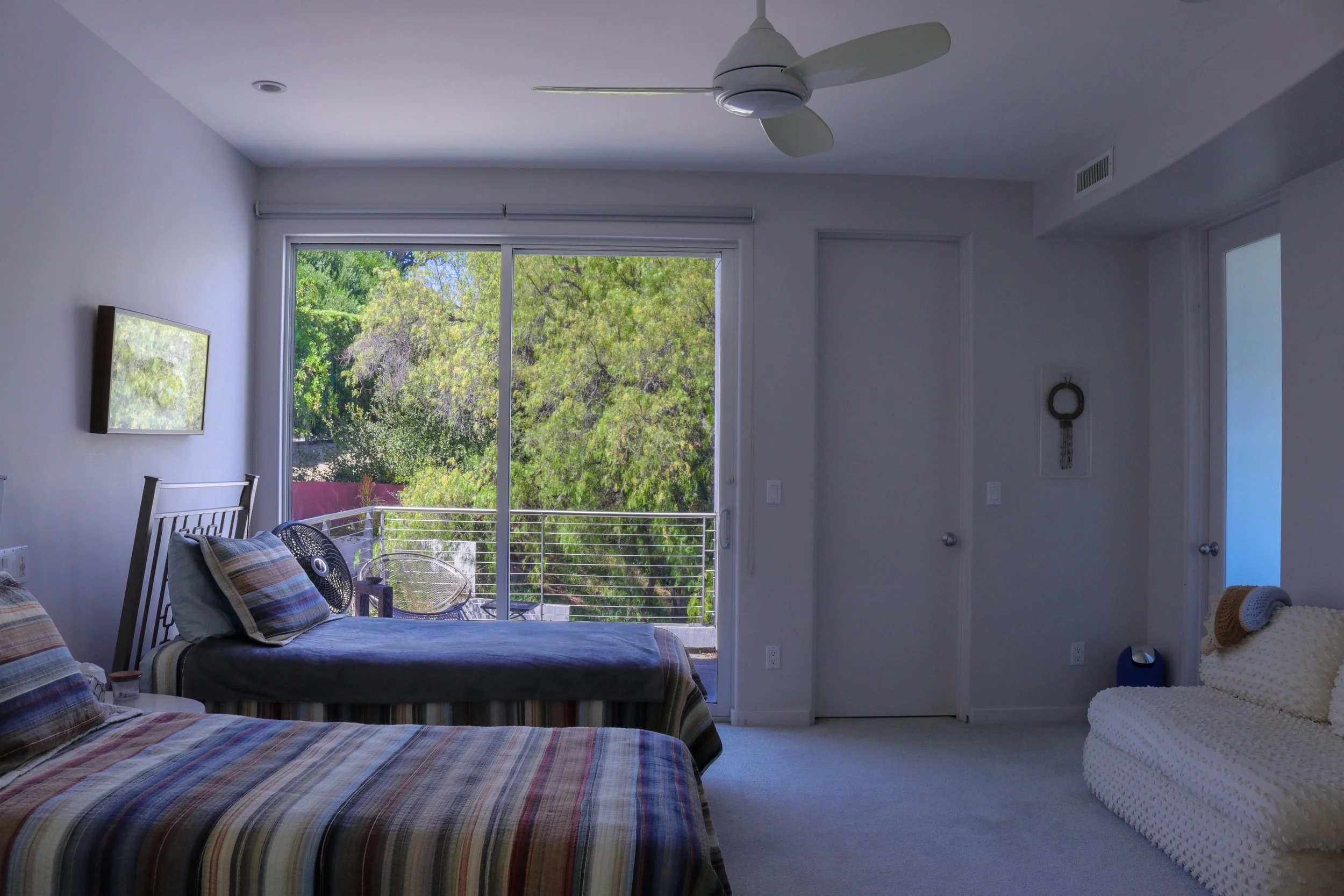 Interior of a bedroom with white walls, a large window showing green trees outside, a bed with striped bedding and pillows, a small black fan, a wall-mounted TV, a balcony with a railing, and a ceiling fan.