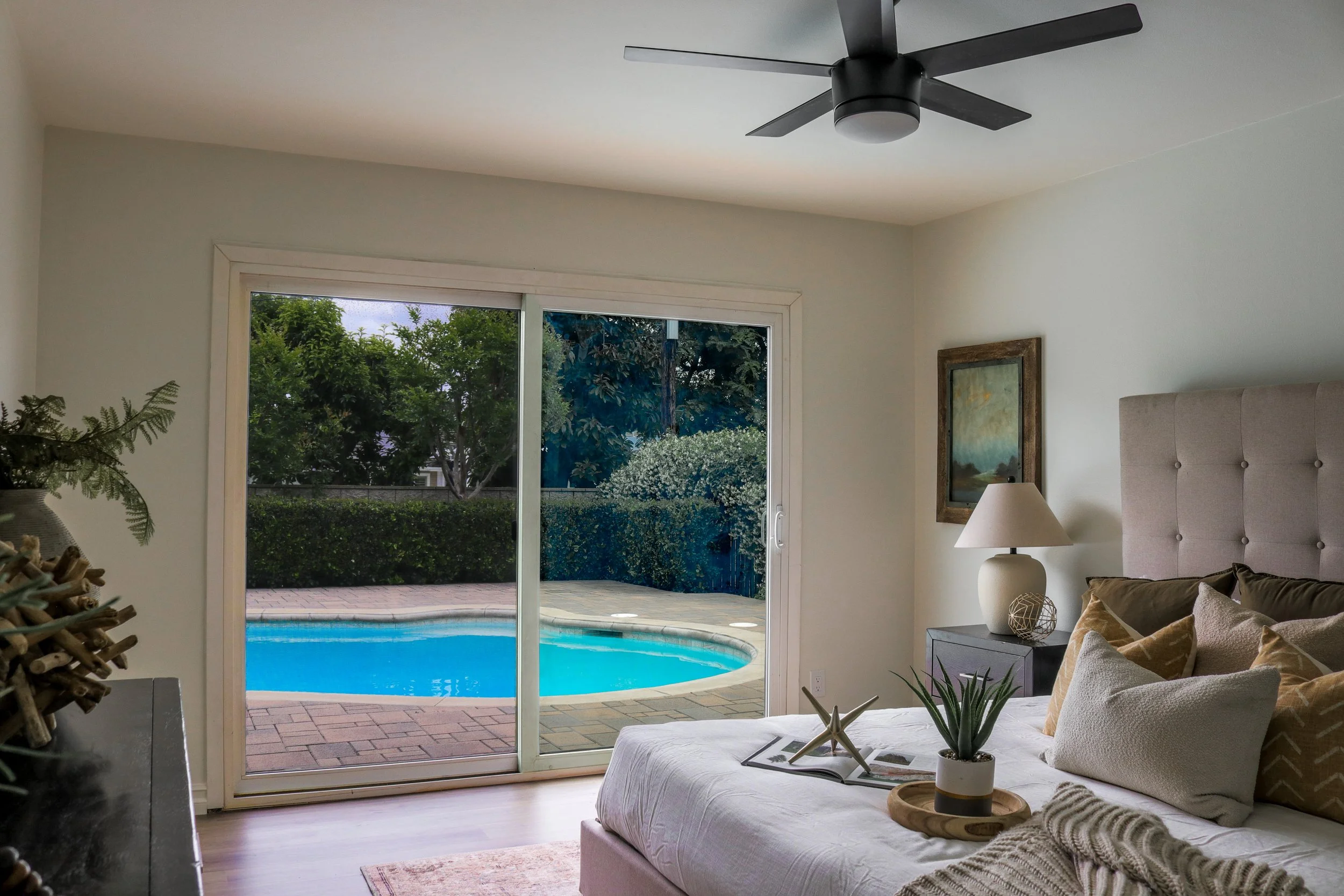 Living room with large sliding glass doors leading to a backyard pool area, a beige sofa with pillows, a side table with a lamp, and decorative plants and artwork.