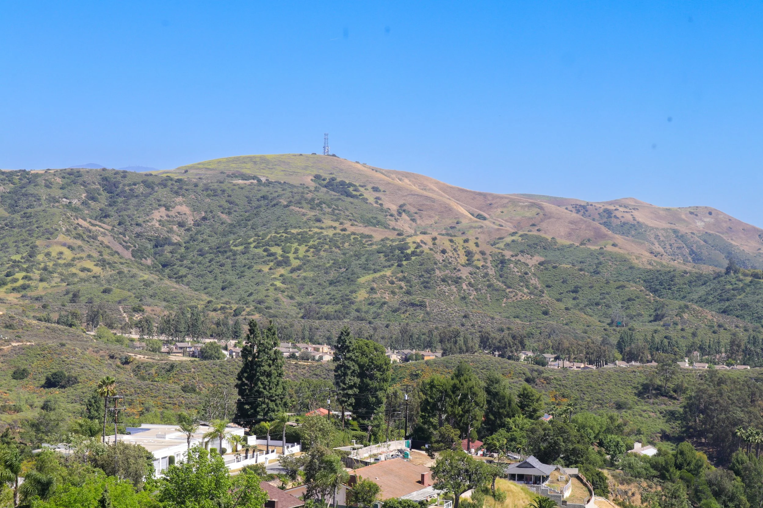 A scenic view of green hills with houses at the base and a communication tower on the hilltop under a clear blue sky.