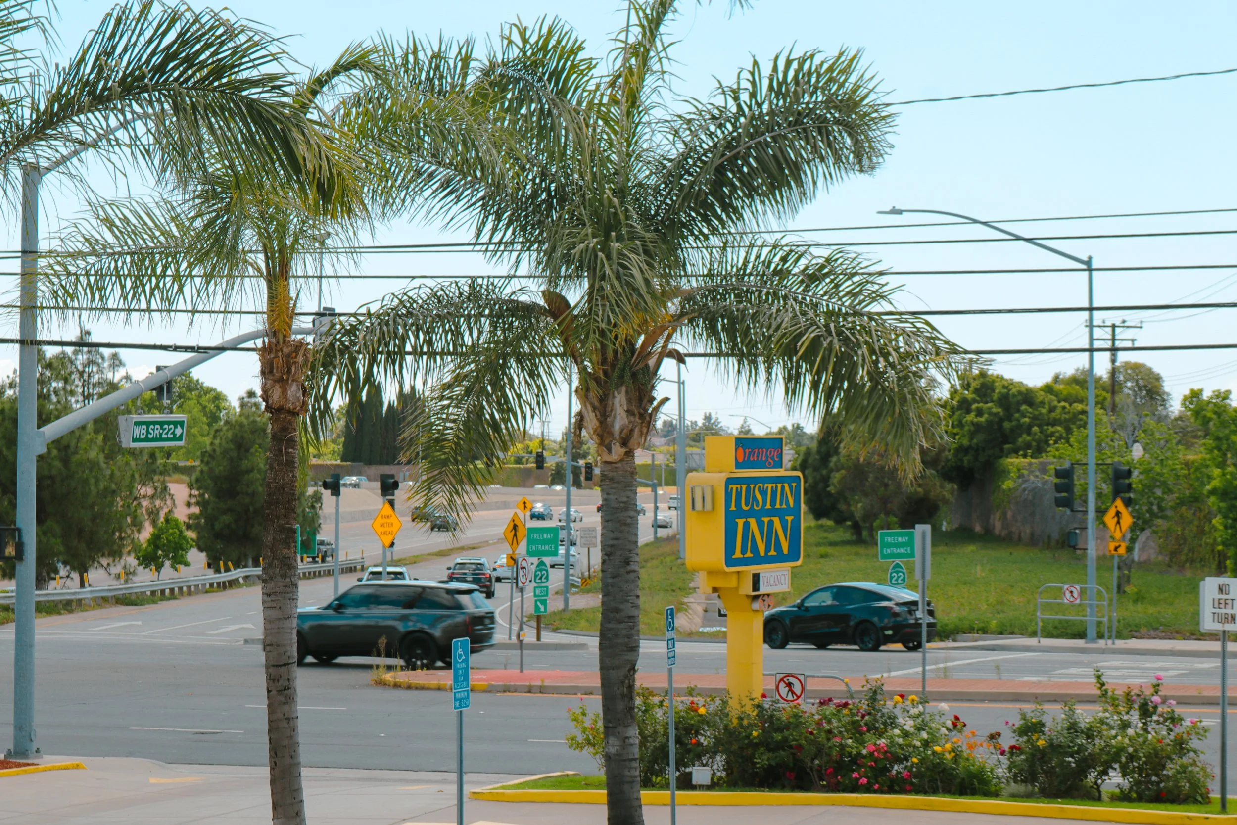 A parking lot with palm trees, traffic signs, and a yellow sign for Tustin Inn. Vehicles are driving on the road, and there are various street signs and a traffic light visible. The sky is clear and blue.