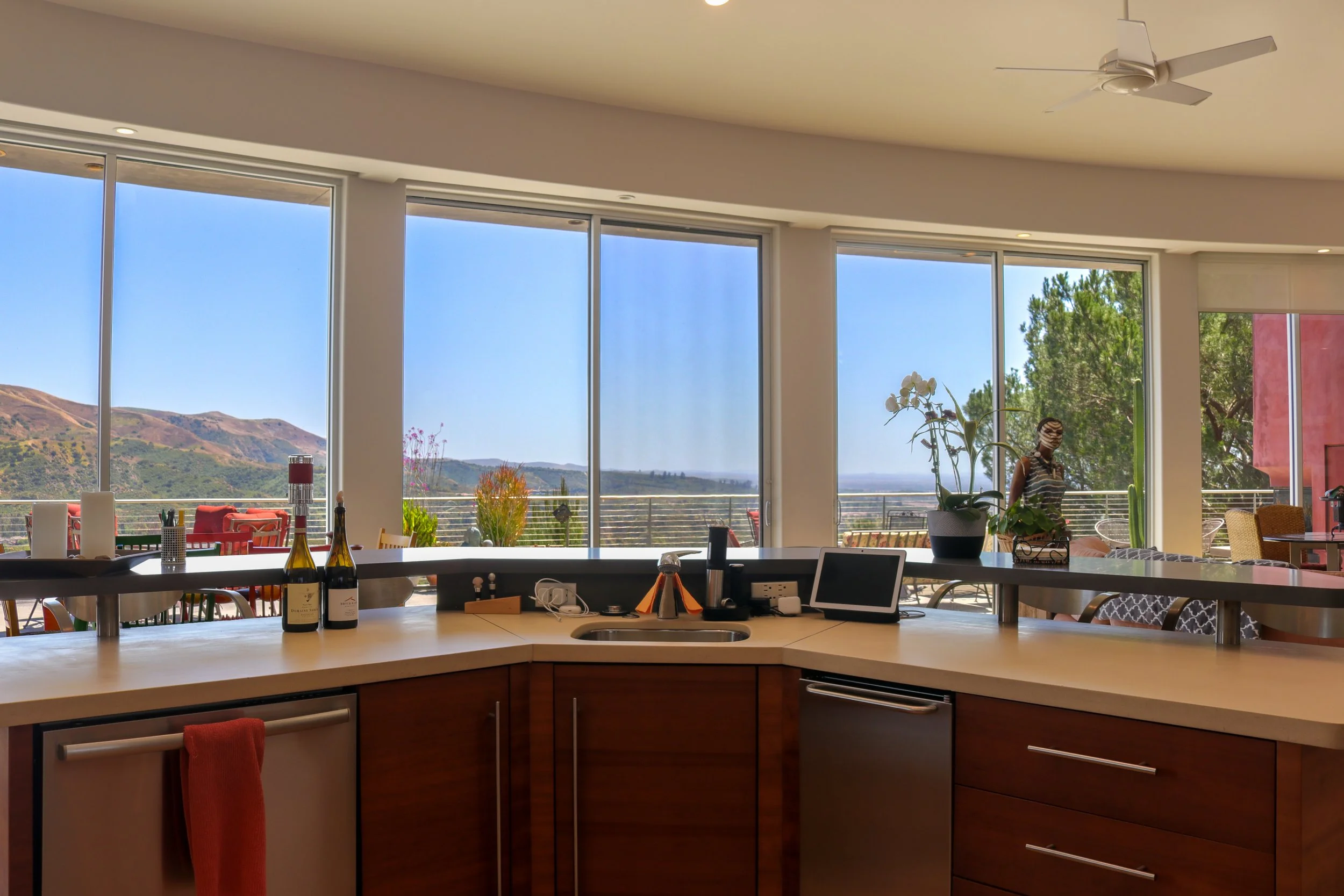 Modern kitchen interior with a large island counter and a view of hills outside through big glass windows. There are bottles, a tablet, and decorative plants on the counter, with a dining and outdoor seating area visible.