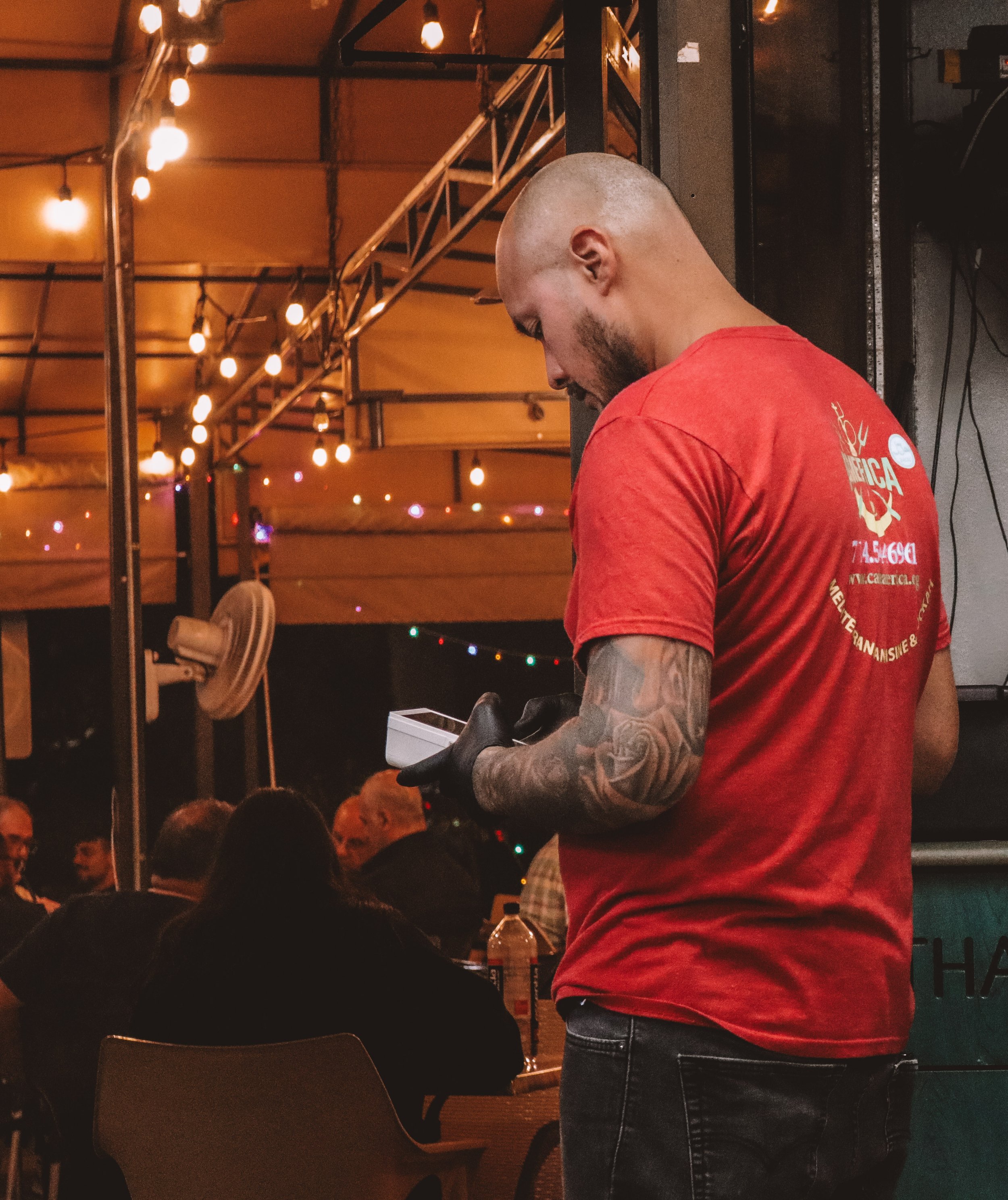 A man with tattoos on his arm, wearing a red shirt and jeans, is looking down at a device in his hands at a dimly lit indoor event with string lights and tables in the background.