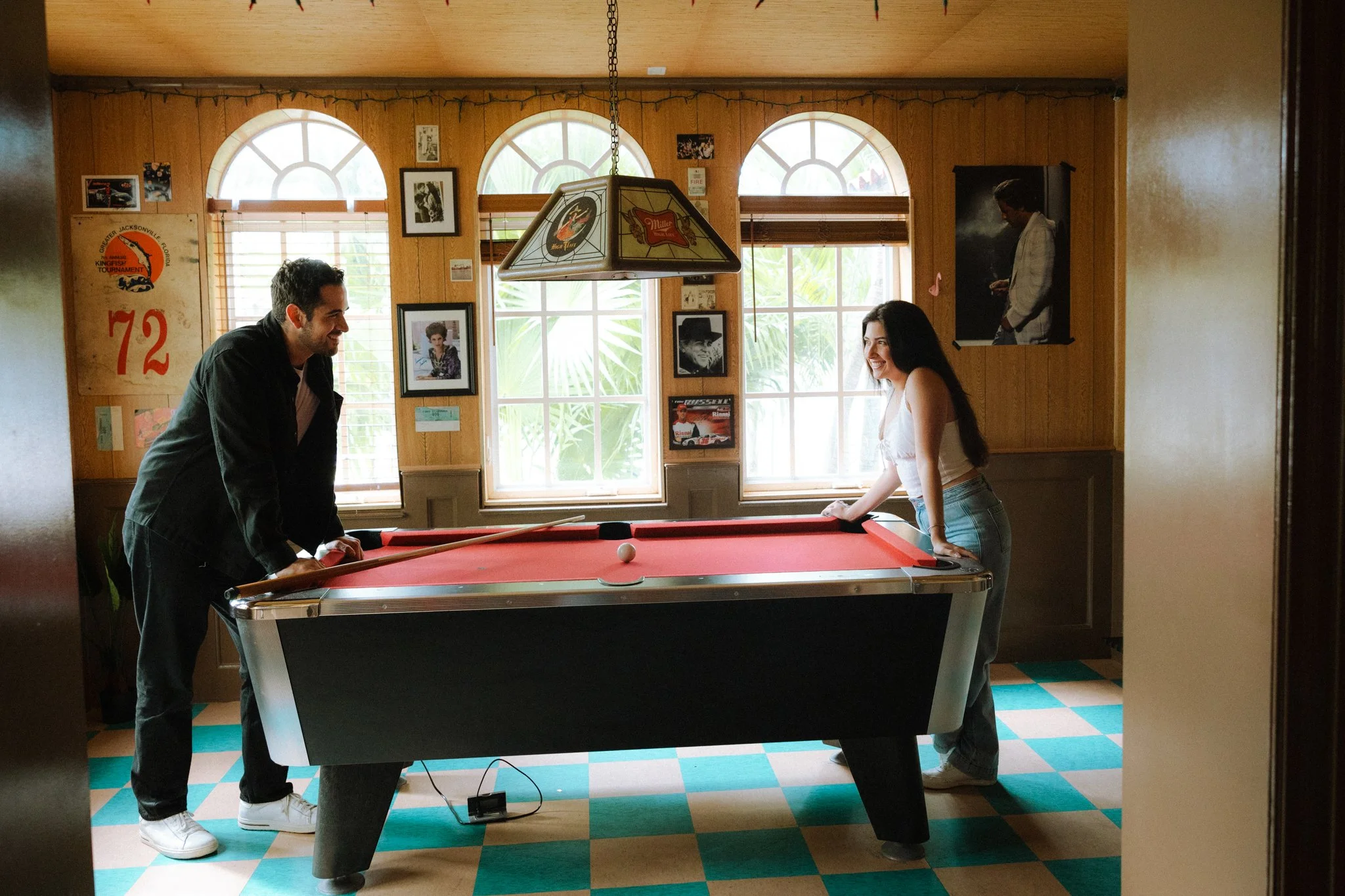 A man and woman playing pool in a room with wooden walls and large arched windows. The man is leaning on the pool table, smiling, and the woman is standing on the other side, also smiling. There are pictures and posters on the walls, including a larg