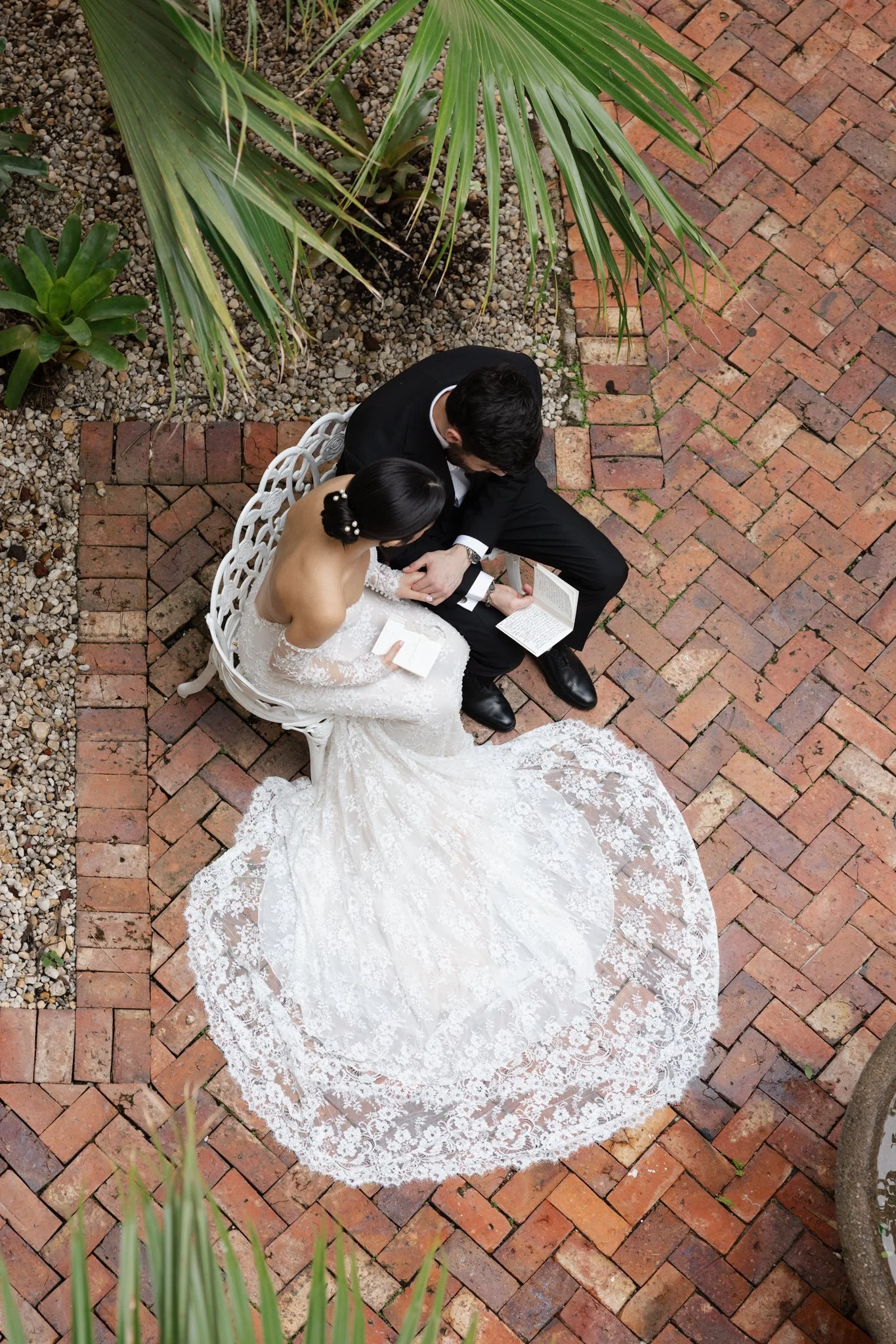 A bride and groom sitting on a white bench with brick flooring and greenery around them, reading a book.