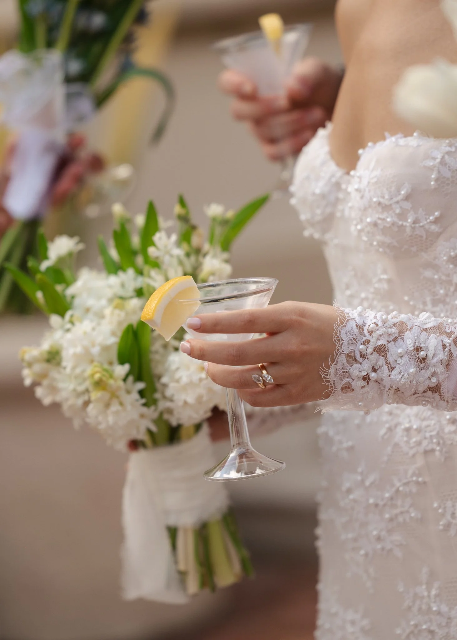 A bride in a lace wedding dress holding a cocktail glass with a lemon wedge, with blurred flowers and another person in the background.