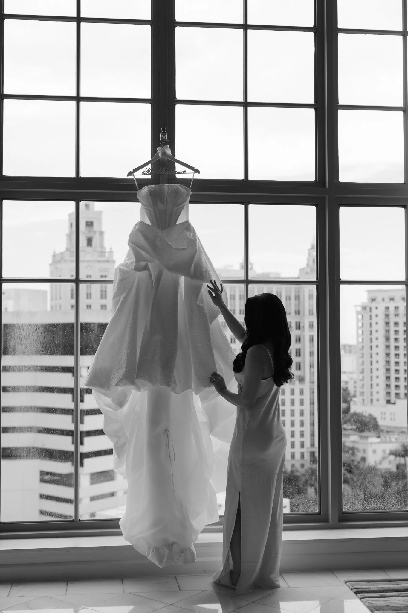 A wedding dress hanging in front of a large window with a city skyline in the background, and a woman reaching up to touch the dress.