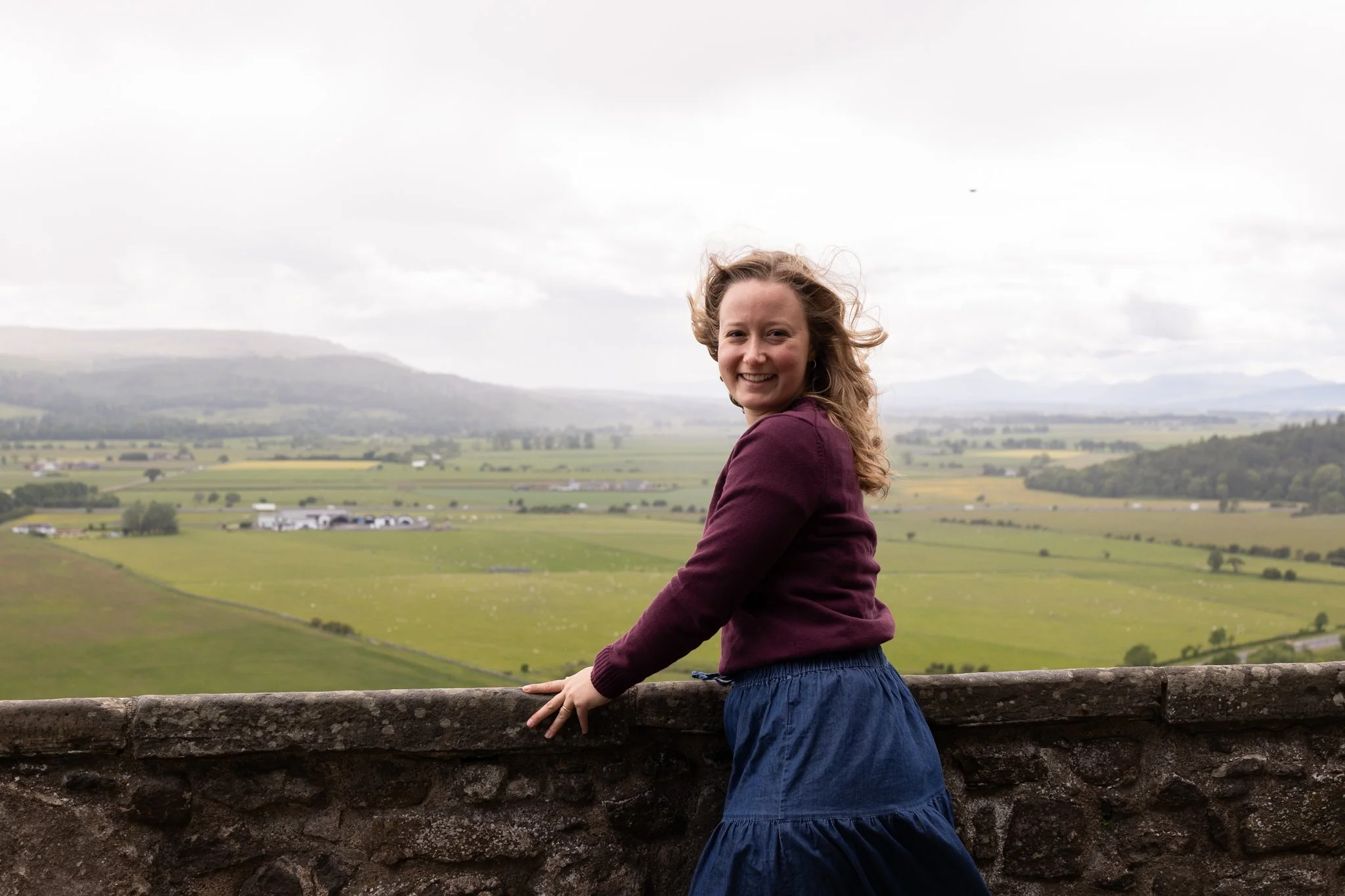 Smiling woman with curly hair wearing a maroon sweater and a blue skirt, standing outdoors on a stone wall with a rural landscape of plains and hills under an overcast sky in the background.