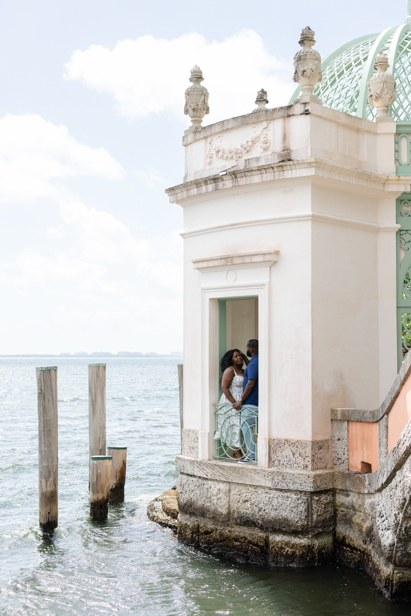 A couple standing on a small balcony by the water, with a historic structure adorned with decorative urns and a glass-domed roof in the background.