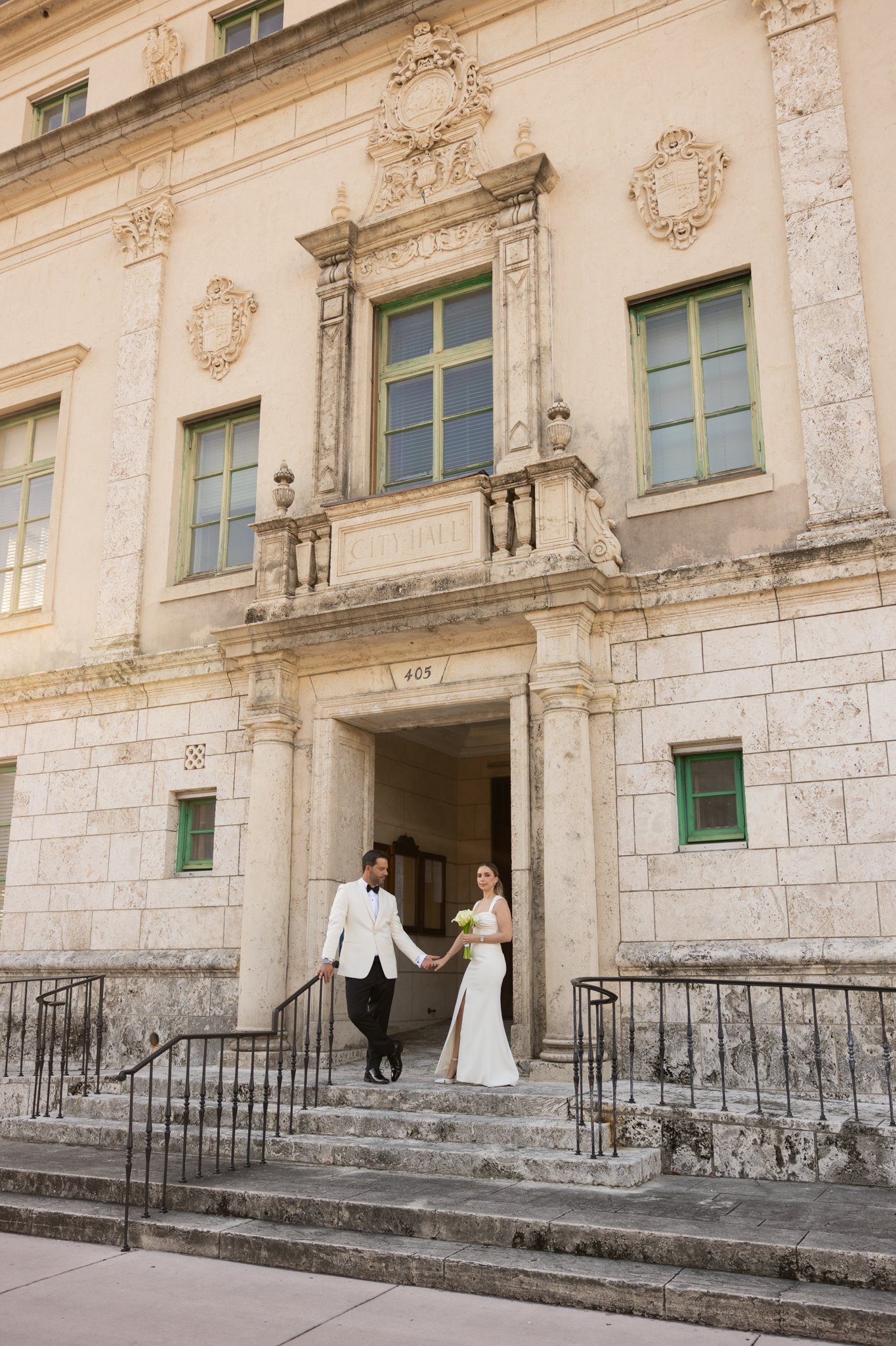 A couple in formal wedding attire standing outside a historic city hall building. The man is in a white tuxedo and the woman in a white wedding dress holding a bouquet of flowers. They are holding hands on the steps of the building.