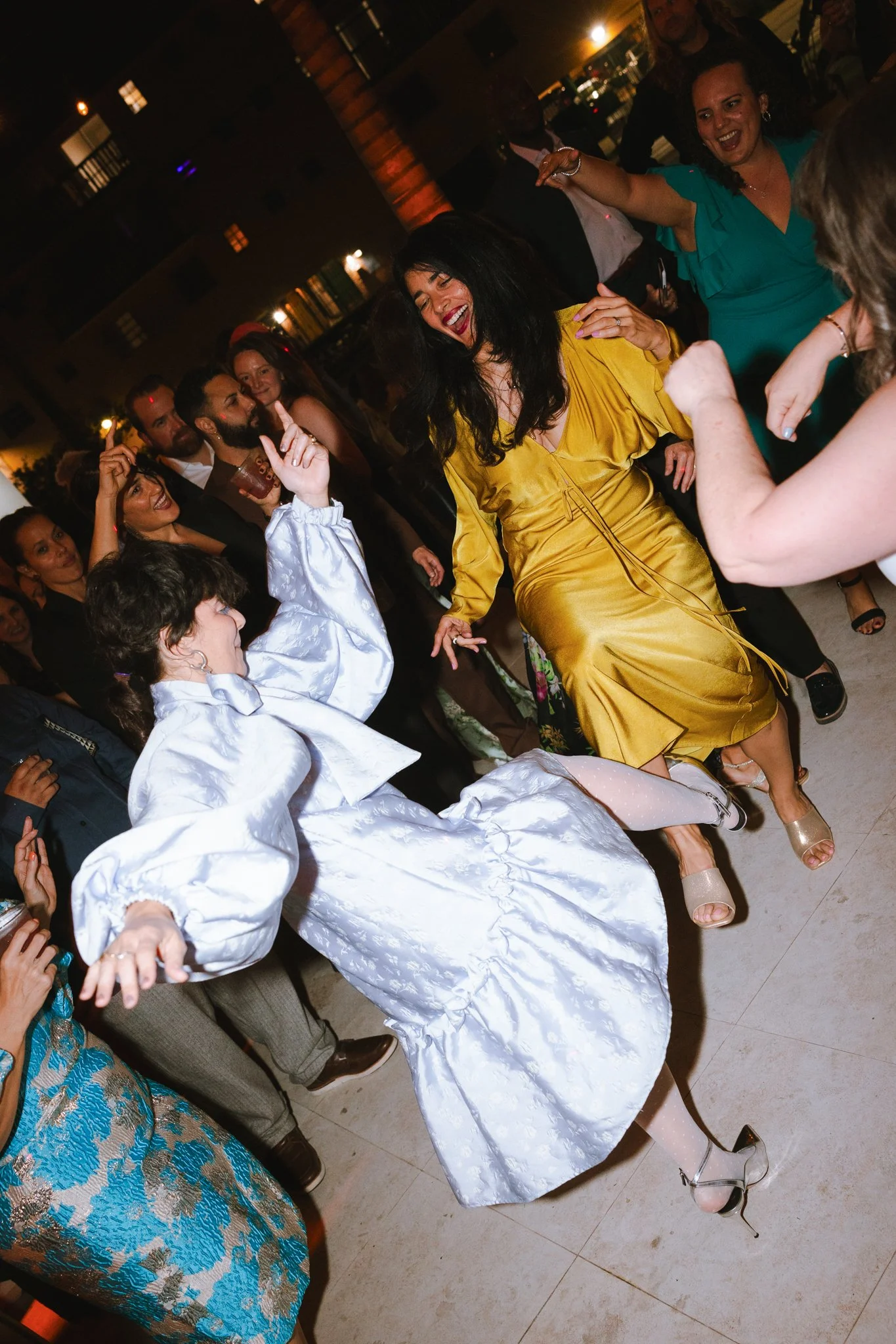 People dancing and enjoying themselves at a party or celebration, with women in colorful dresses, including a woman in a yellow satin dress and a woman in a teal dress, in a lively indoor setting.