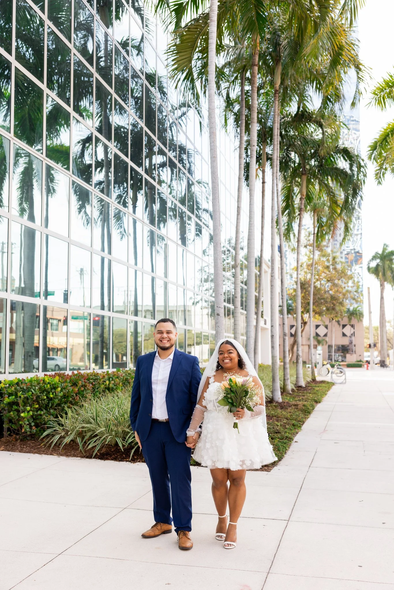 A joyful couple walks hand in hand outdoors along a sidewalk, with tall palm trees and a modern glass building reflecting trees in the background. The man is dressed in a blue suit, white shirt, and brown shoes, and the woman in a white wedding dress holding a bouquet, wearing white heels. They are smiling happily.
