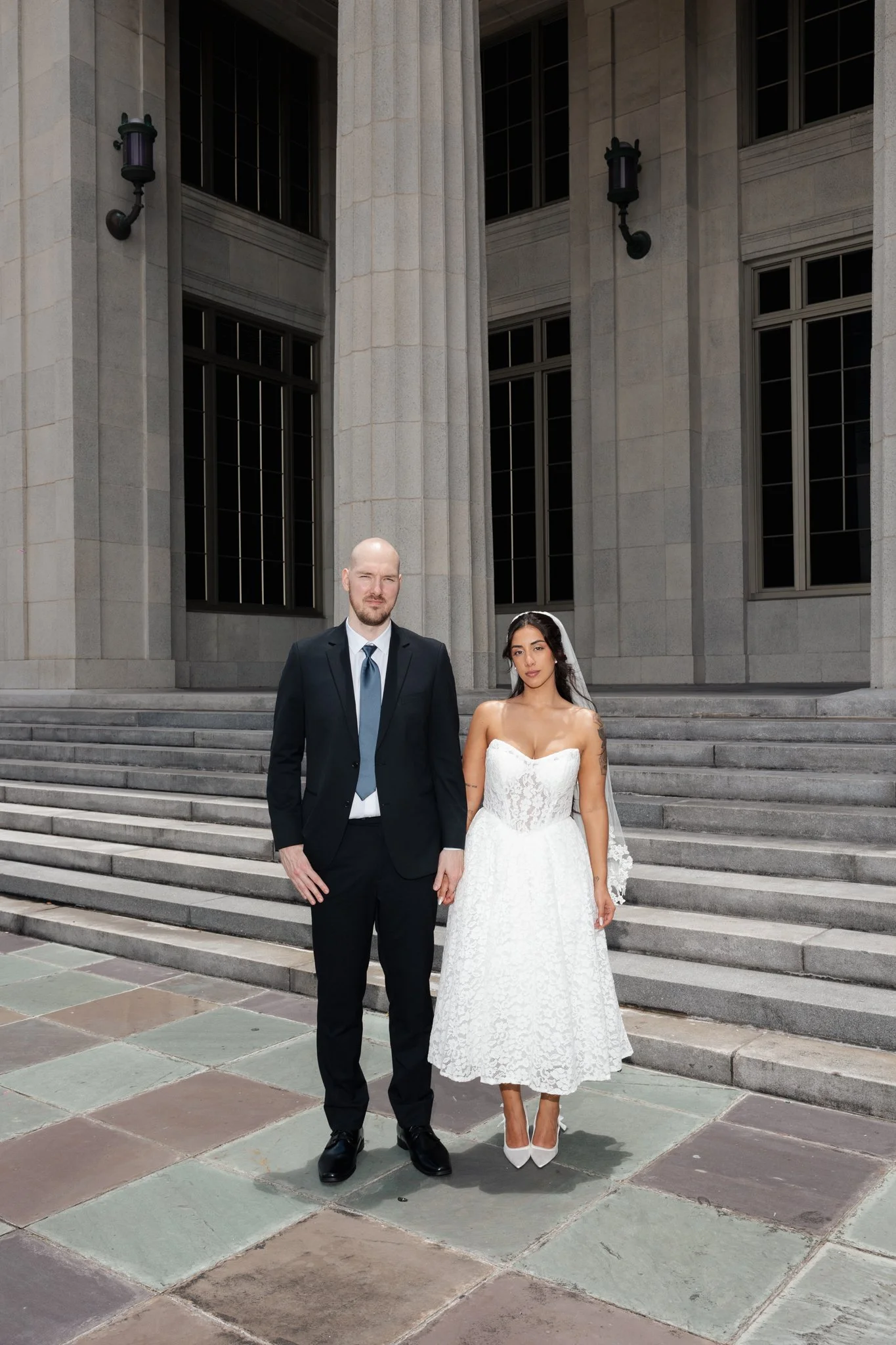 A bride and groom stand on steps outside a stone building with large windows and decorative lights.