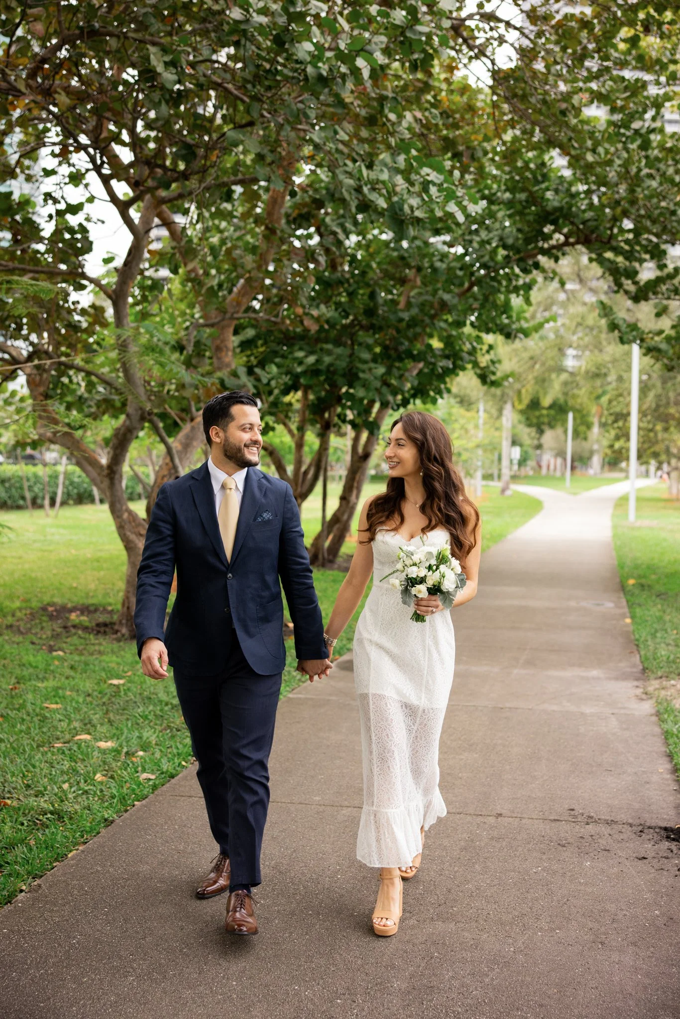 A man and woman dressed in wedding attire walking hand in hand along a park sidewalk, smiling at each other, with trees and green grass in the background.