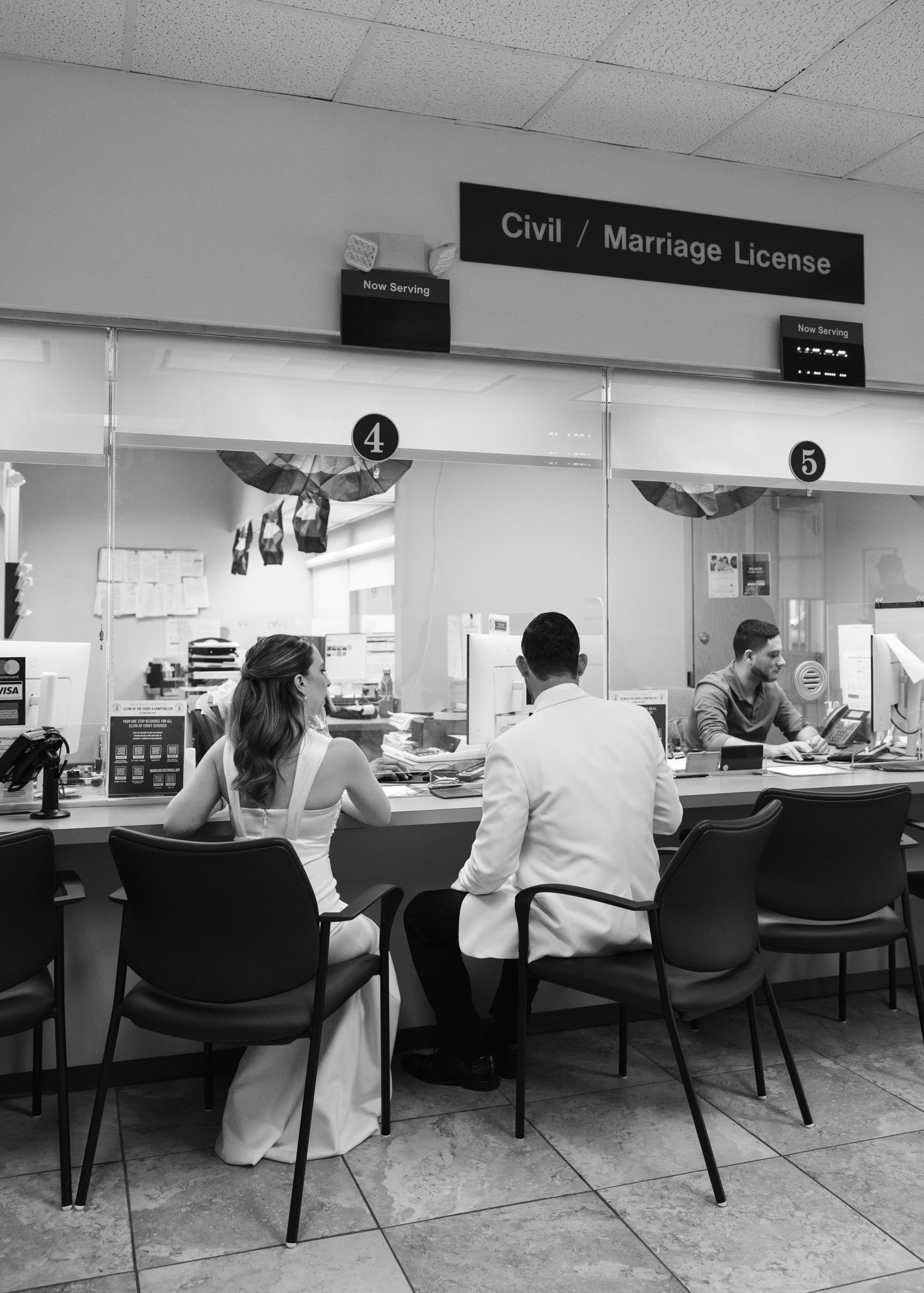 People sitting at a counter in a government office, with signs indicating they are applying for or processing civil or marriage licenses.