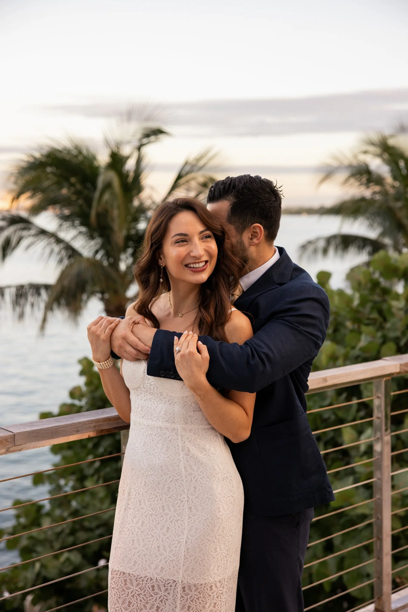 A smiling woman in a white dress embracing a man in a dark suit on a wooden balcony overlooking water with palm trees in the background during sunset.