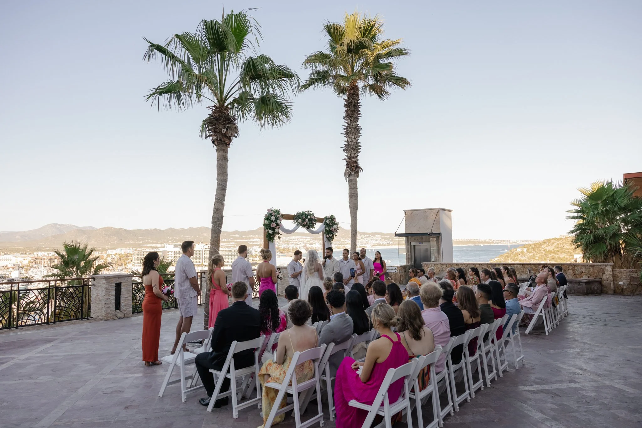 A wedding ceremony taking place outdoors on a terrace with ocean and mountain views, palm trees, and a floral arch. Guests are seated and standing around the couple and officiant.