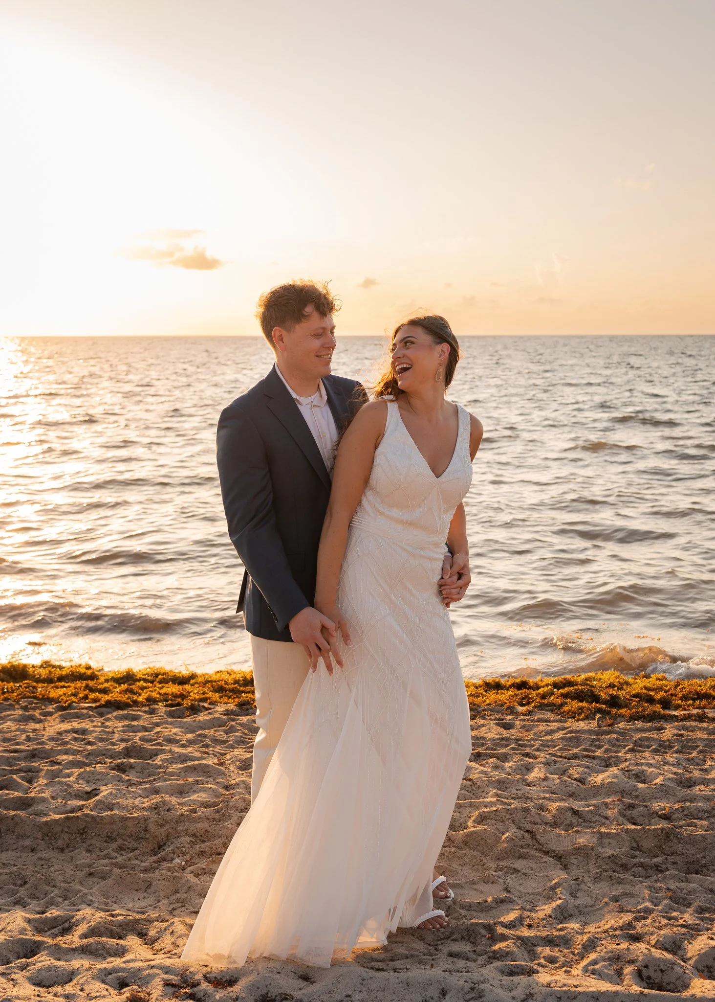 A couple in wedding attire standing on a beach at sunset, smiling and holding hands.