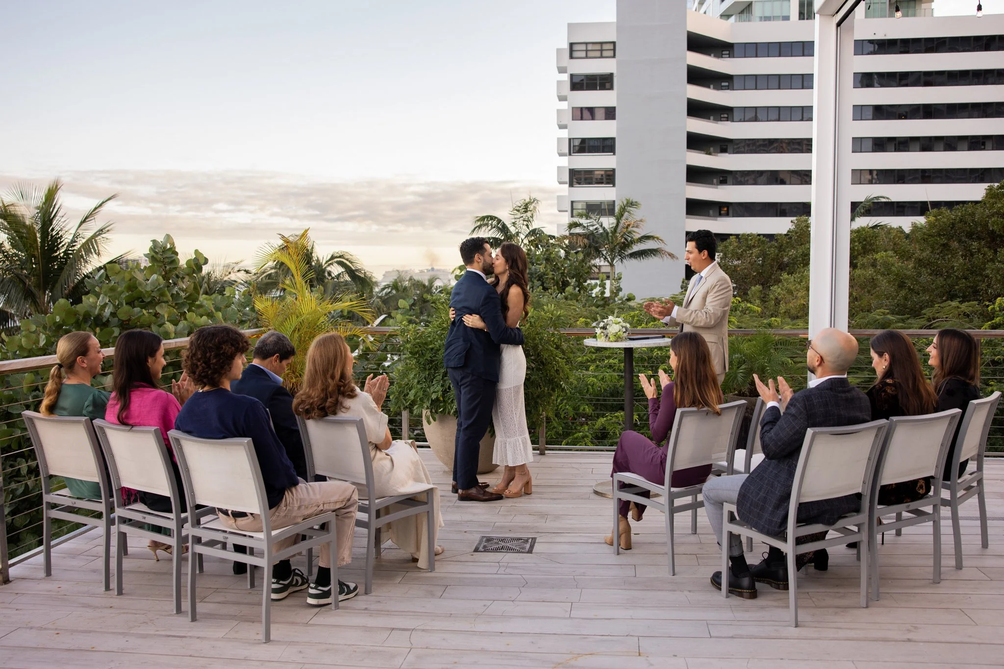 A wedding ceremony on a rooftop terrace with urban buildings in the background. A bride and groom are kissing while an officiant stands nearby. Guests are seated, clapping and smiling. There are green plants and string lights overhead.