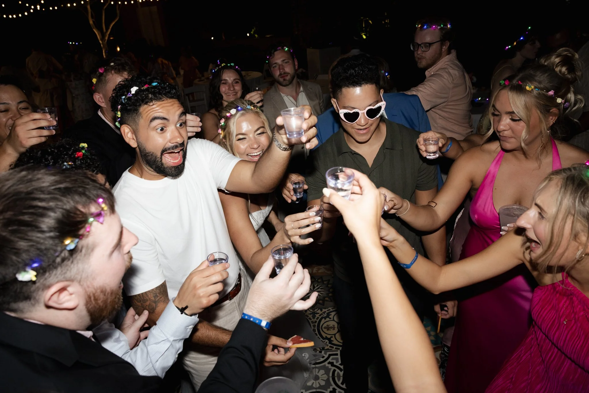 Group of people celebrating at a party, some wearing colorful flower crowns, holding drinks, with smiles and joyful expressions, in a dimly lit setting with party accessories.