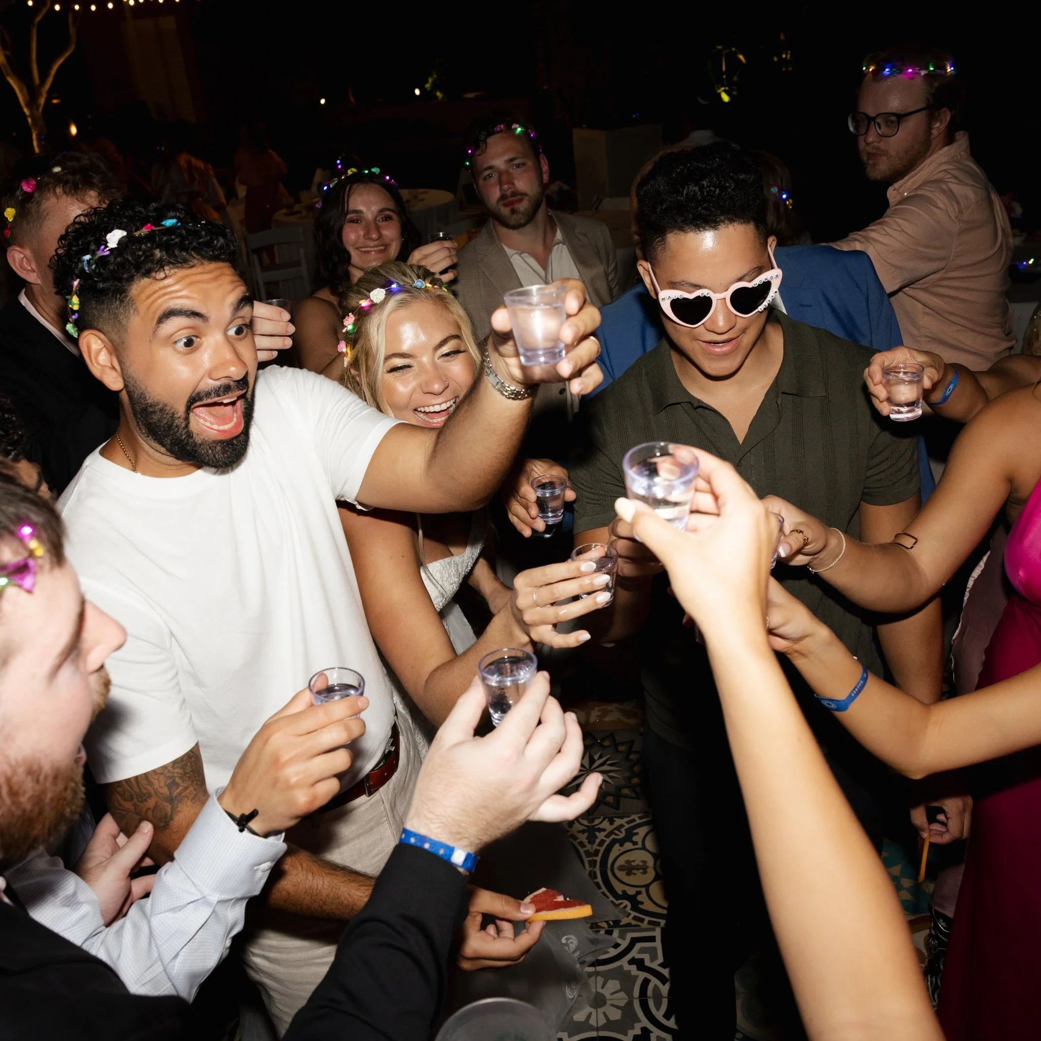 A group of people celebrating at a party, wearing colorful flower crowns, holding drinks, and smiling in a dimly lit room.