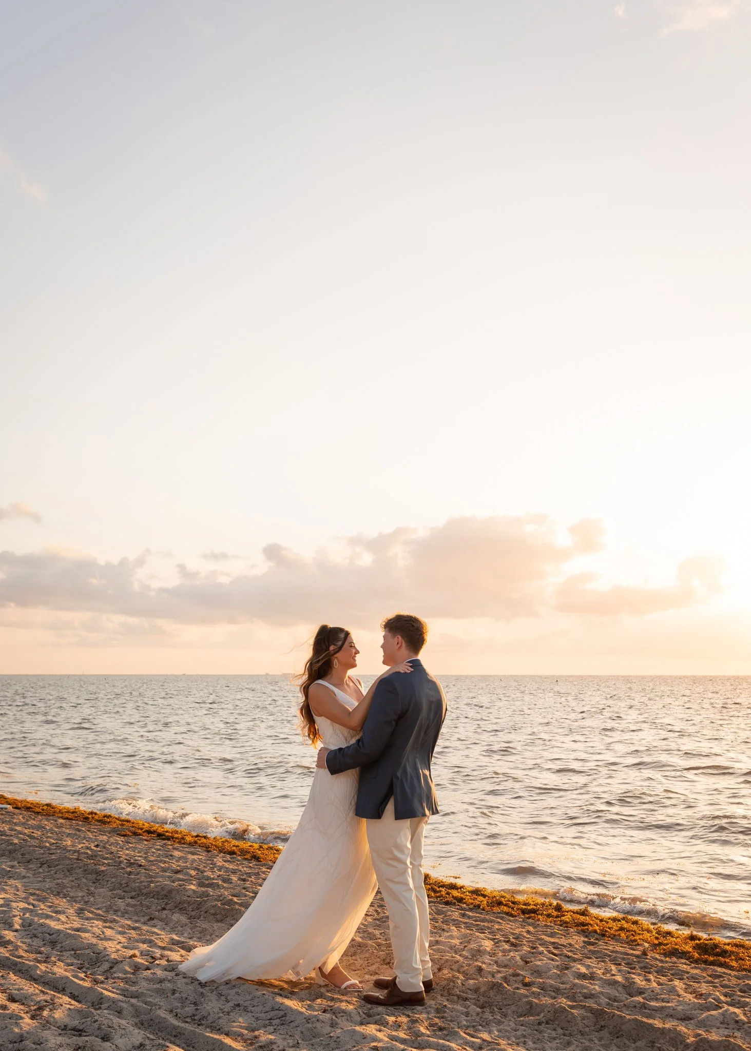 A couple in wedding attire stands on the beach at sunset, facing each other and holding close, with the ocean and a cloudy sky in the background.