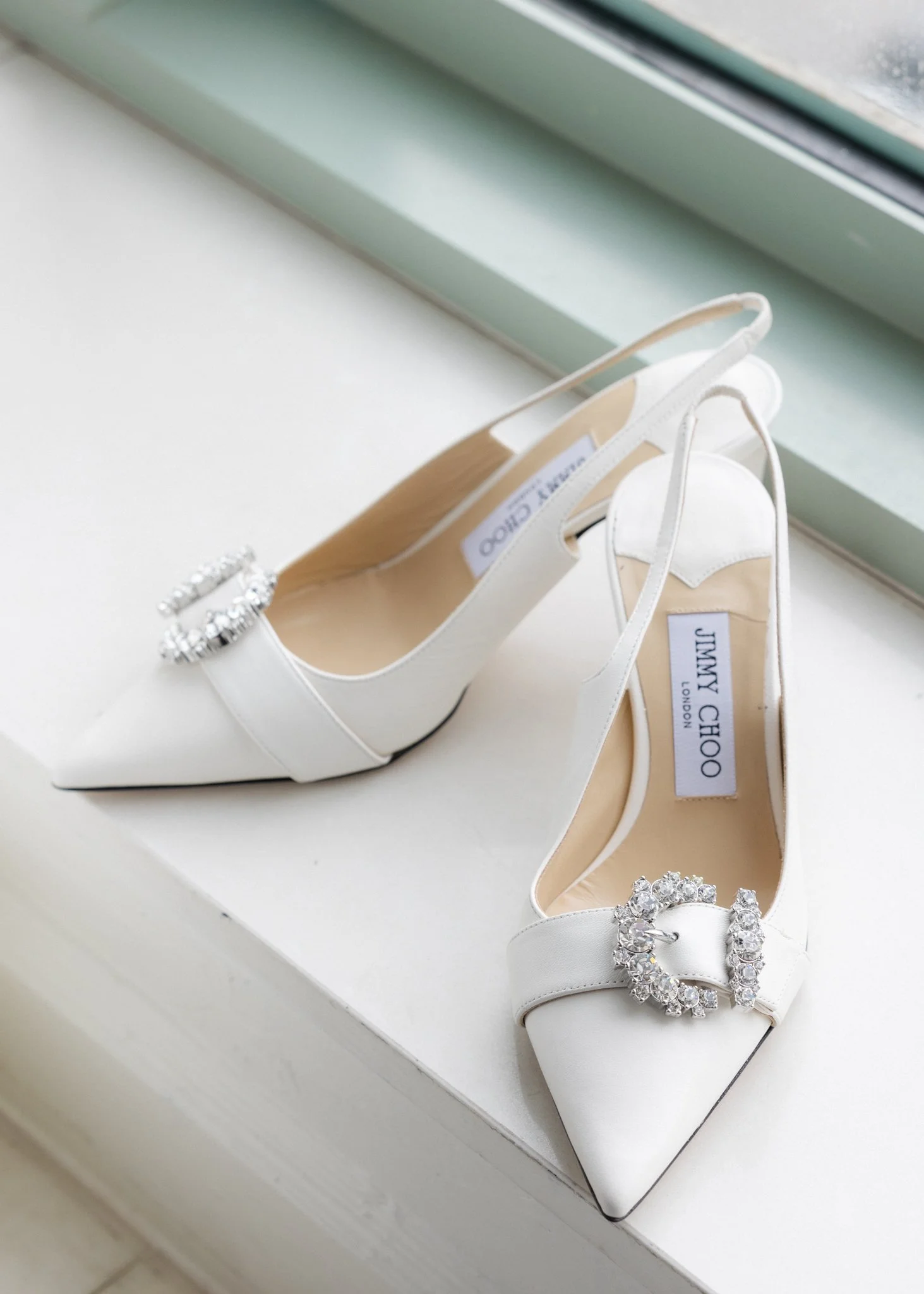White high-heeled shoes with rhinestone embellishments on the toes, placed inside white shoeboxes on a white windowsill.