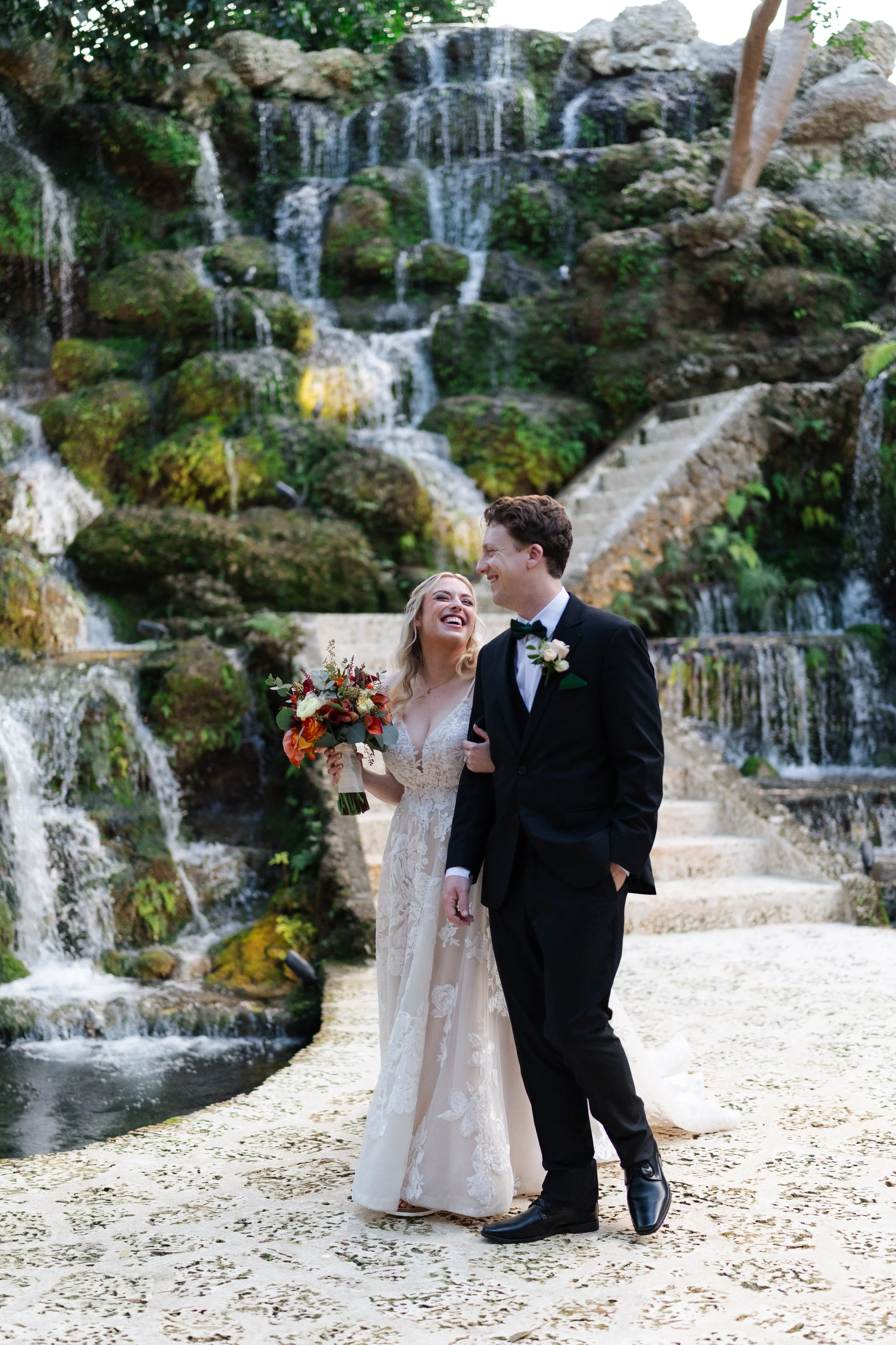 A bride and groom standing together in front of a multi-tiered waterfall with lush greenery. The bride is holding a bouquet of flowers and wearing a lace wedding dress, while the groom is in a black tuxedo with a bow tie.