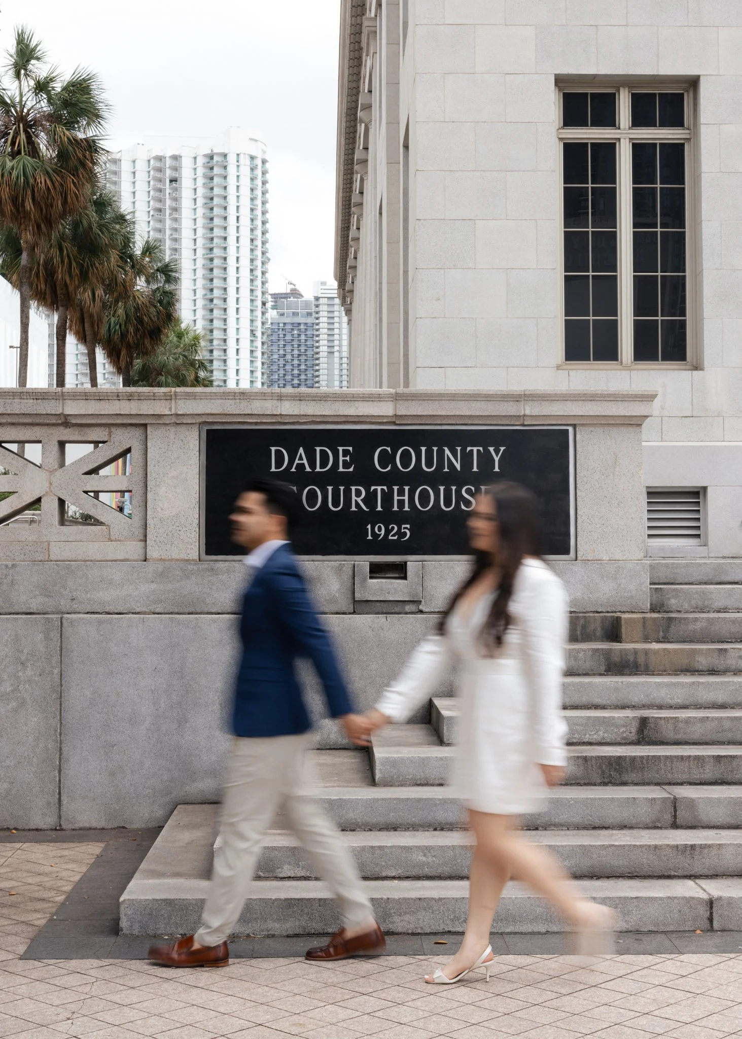 People in formal attire holding hands and walking down steps in front of a courthouse with a sign reading "Dade County Courthouse 1925". Tall buildings and palm trees are in the background.