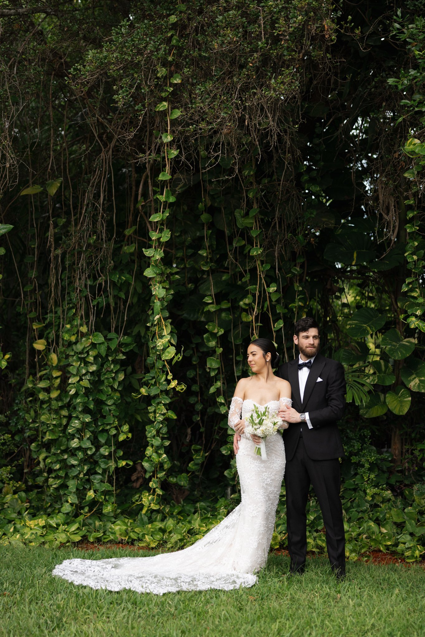 A bride and groom standing outdoors in front of lush green foliage, with the bride holding a bouquet of white flowers and the groom dressed in a black tuxedo, both looking off to the side.