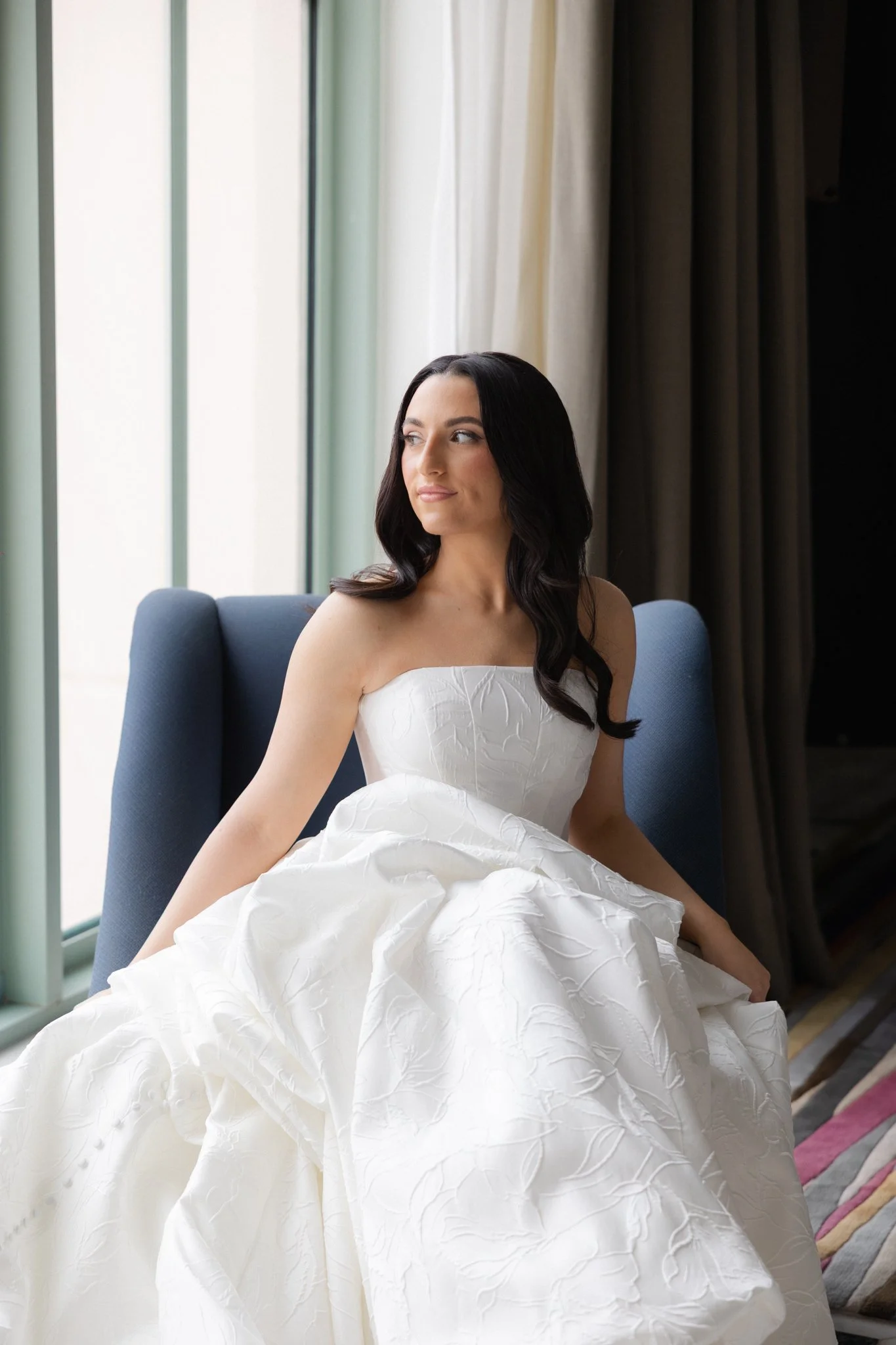 A woman with long black hair, wearing a strapless white wedding dress, sitting on a blue chair near a window with light-colored curtains, looking to the side.