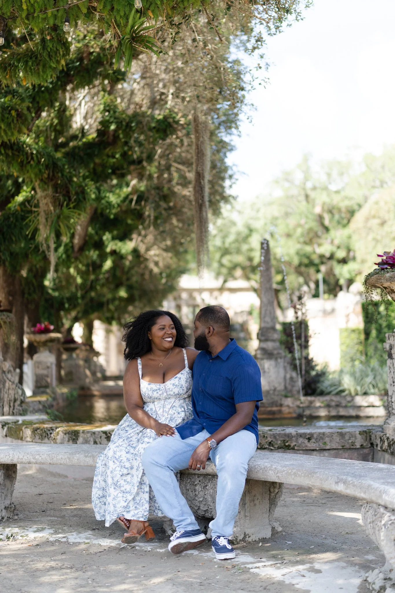 A smiling woman and a man sitting on a stone bench outdoors under trees, engaged in conversation, with lush greenery and a fountain behind them.