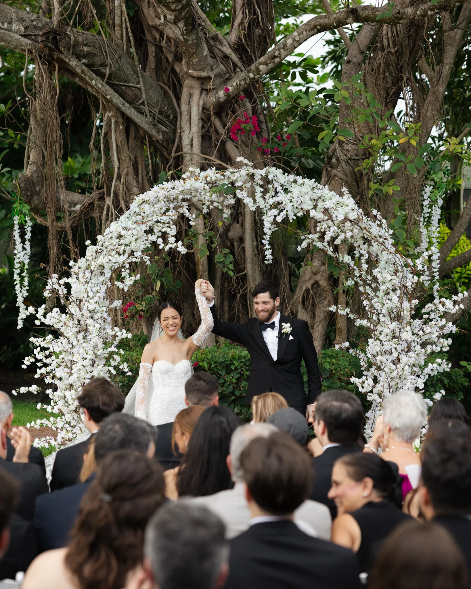 A bride and groom standing under an arch of white flowers, celebrating their wedding, surrounded by guests.