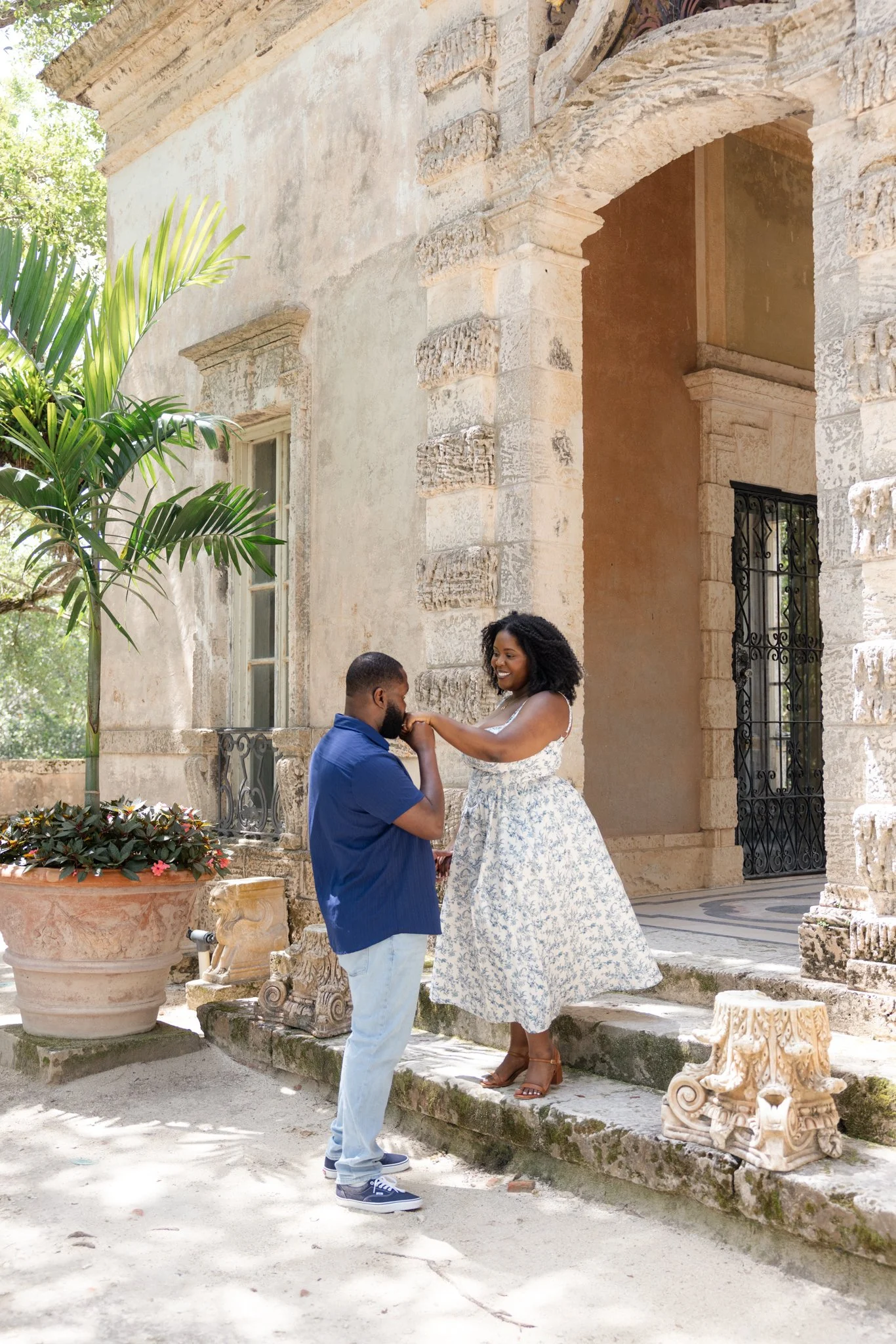 A couple is kissing in a romantic pose outside a historic stone building with decorative columns and iron window grills. The woman is wearing a white floral dress and high heels, while the man is wearing a blue shirt and light jeans. There are potted