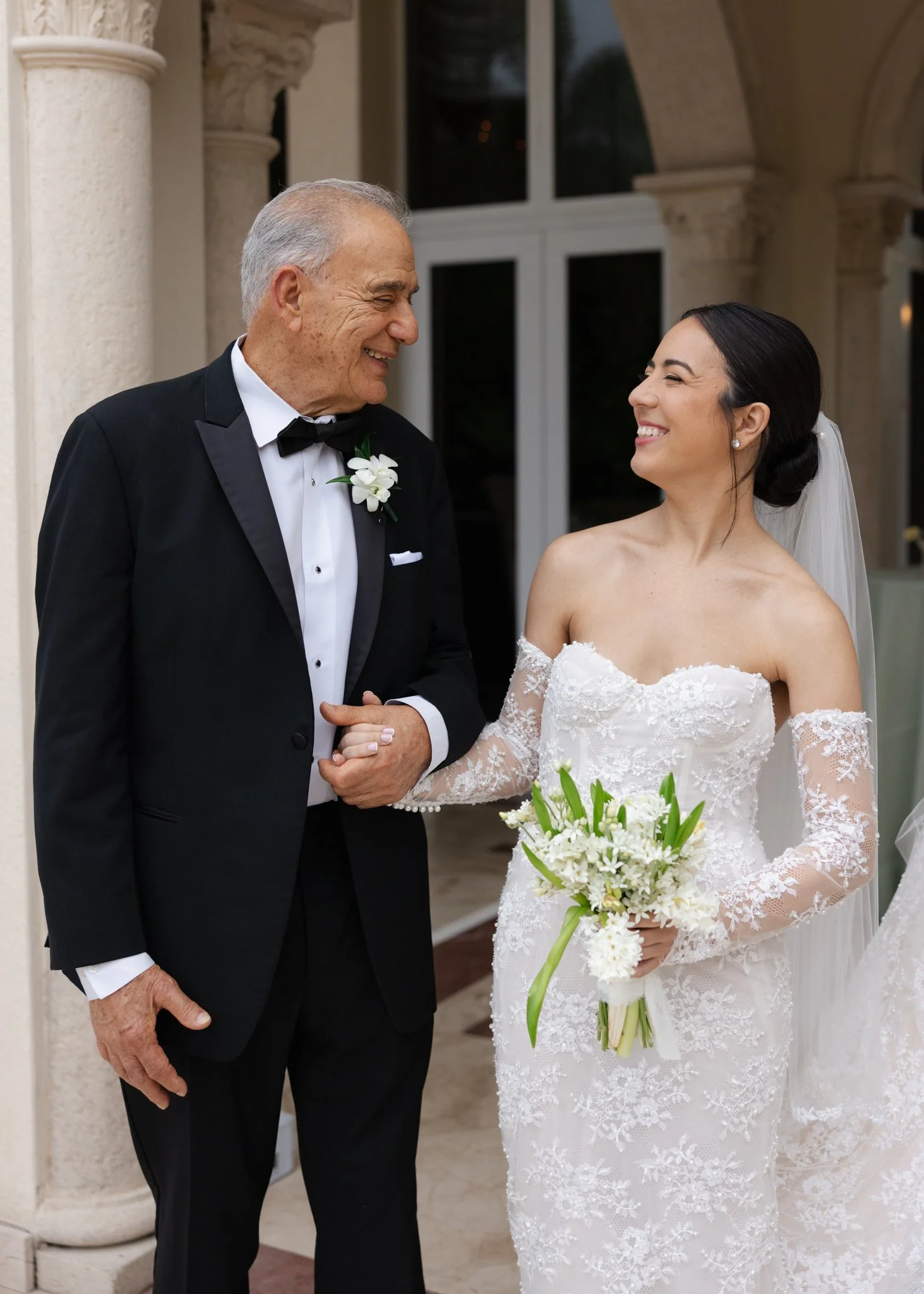 An elderly man and a young woman smiling at each other, dressed in formal wedding attire, standing in front of a building with columns.
