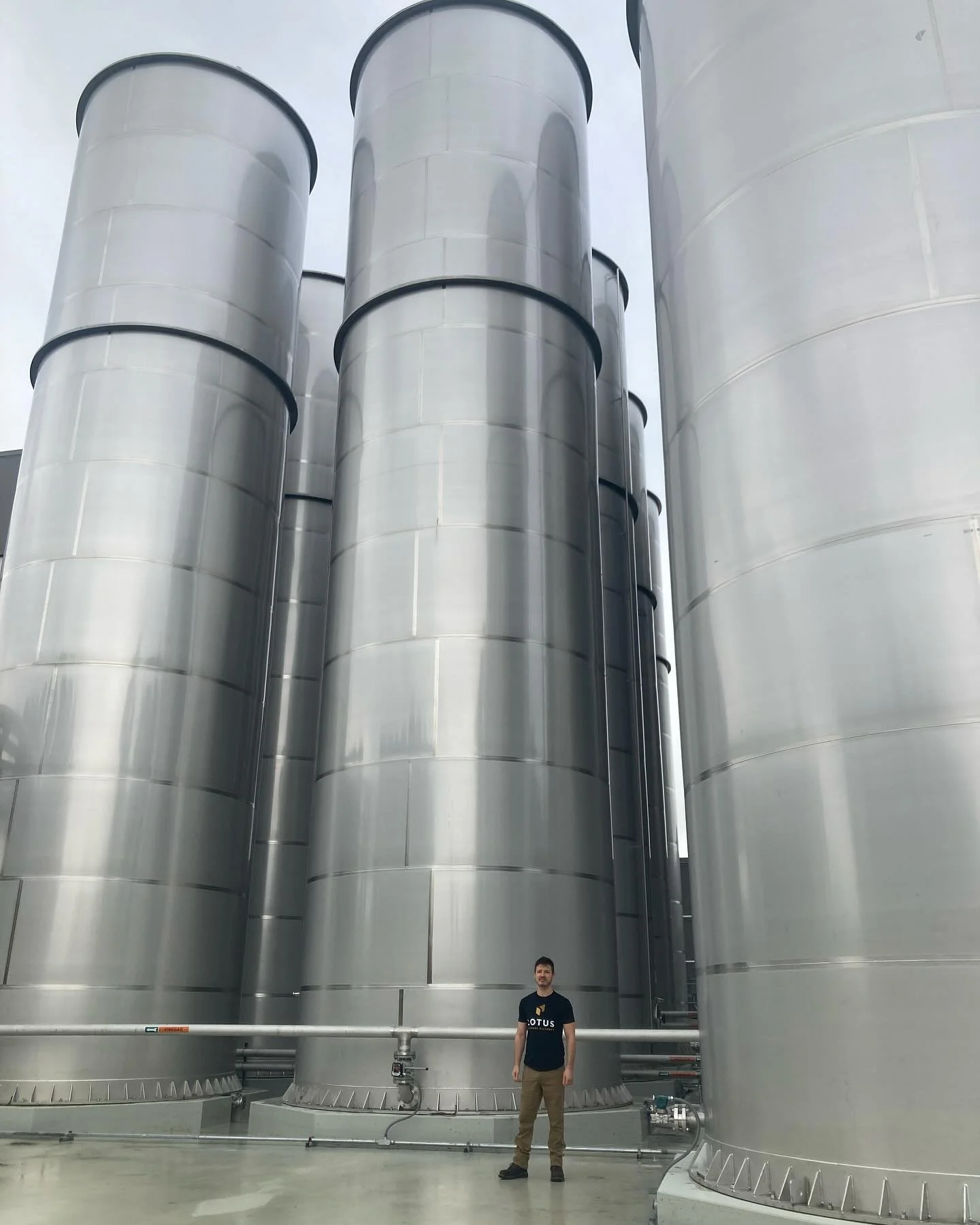 A man standing in front of four large, vertical, metallic industrial storage tanks or silos.