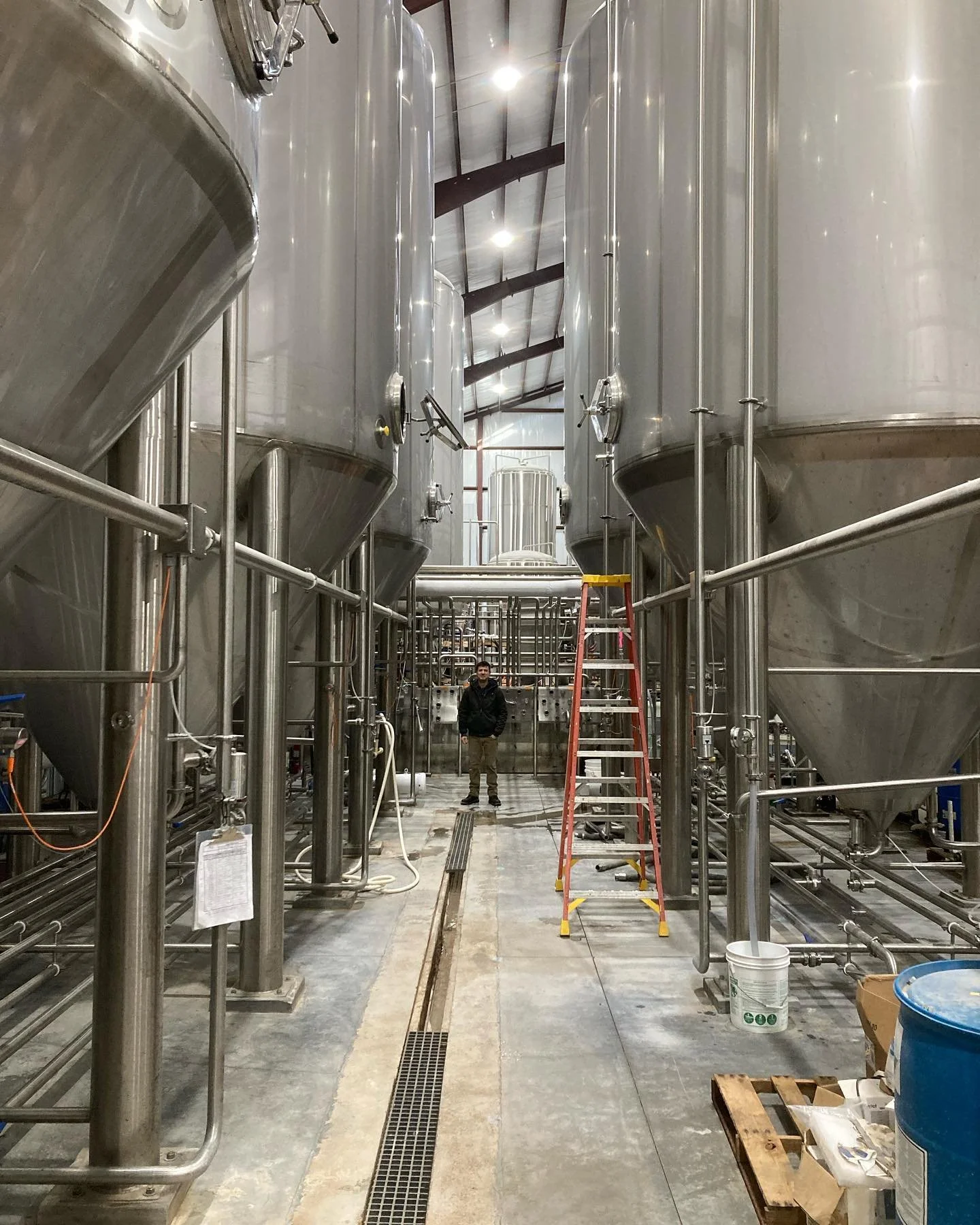 Industrial brewing facility with large stainless steel fermentation tanks, a person standing in the center, and a ladder in the foreground.