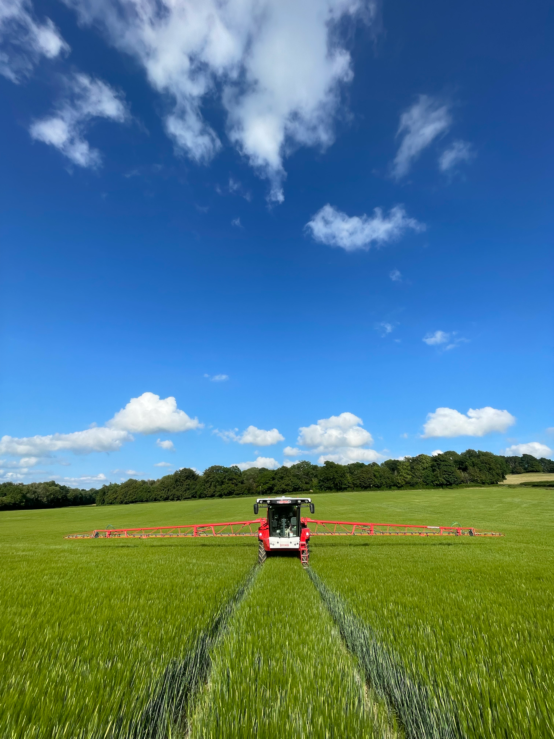 A large agricultural sprayer in a green field under a blue sky with clouds, spraying liquid on the crops.
