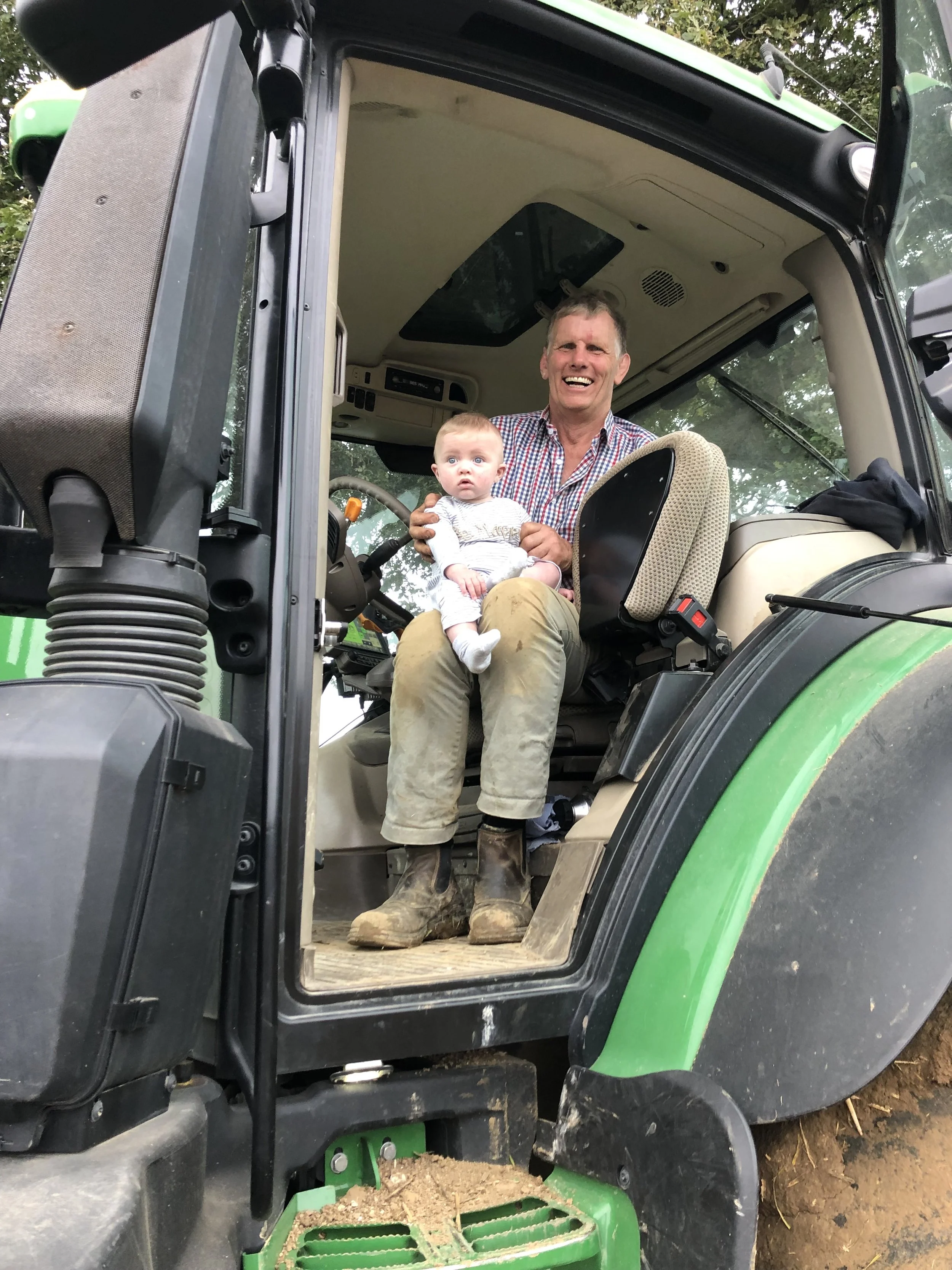 A man and a young child sitting inside a large green tractor, with the man smiling and the child looking surprised, both dressed in casual clothes and boots, with the tractor's front window and dashboard visible.