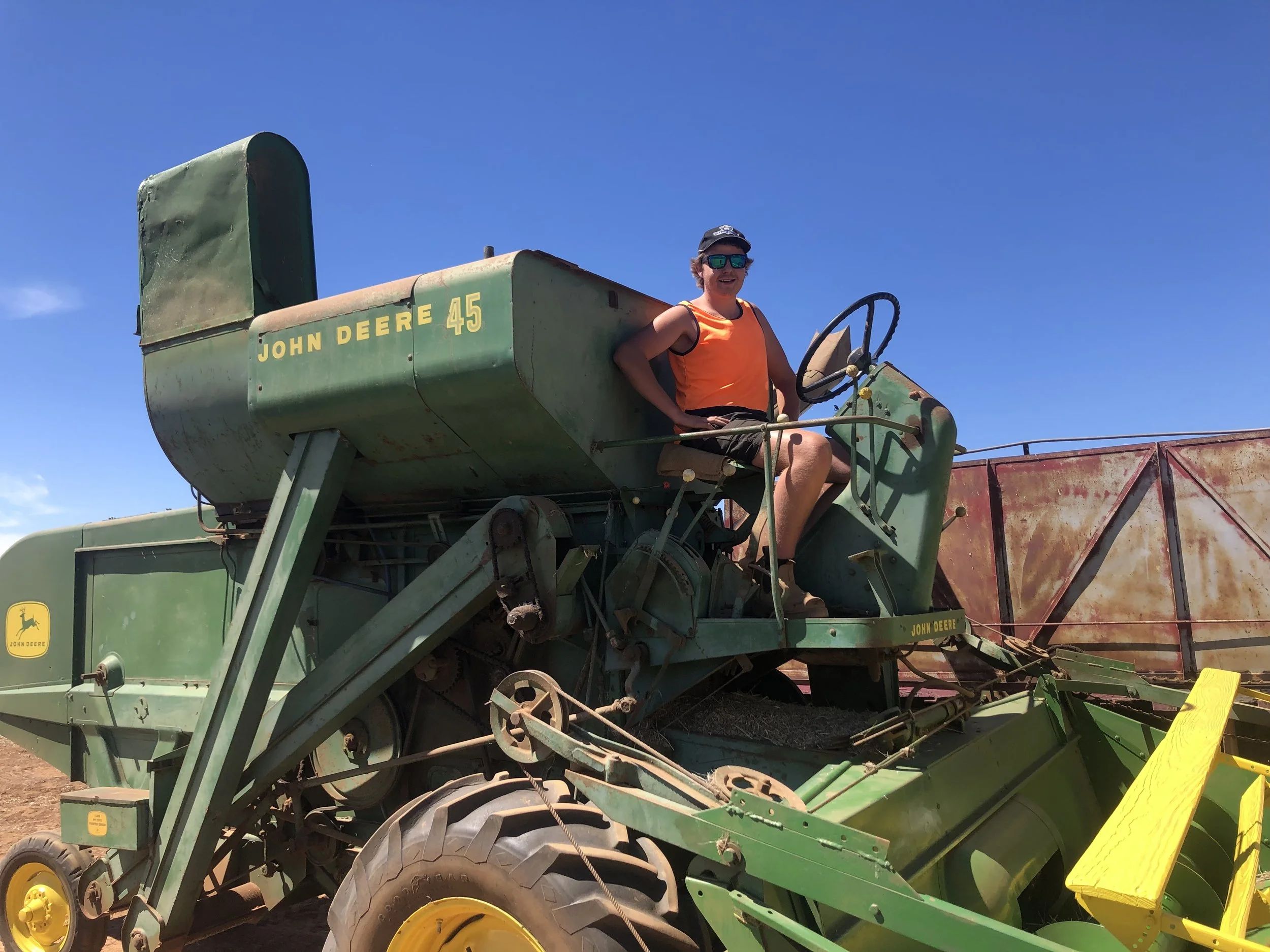 A young man wearing sunglasses, an orange tank top, and a cap sitting on a green vintage John Deere tractor against a clear blue sky.